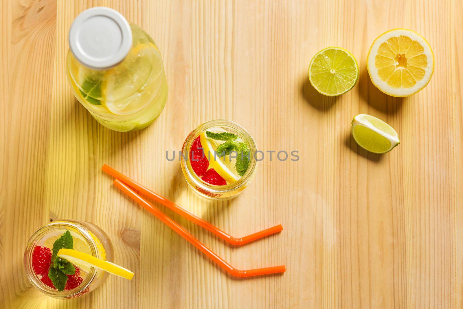 top view, two glass jars and a bottle with refreshing cold lemonade water, lemon, red berries and mint on a wooden table with pieces of citrus lit by sunlight, Summer refreshment background Copy space