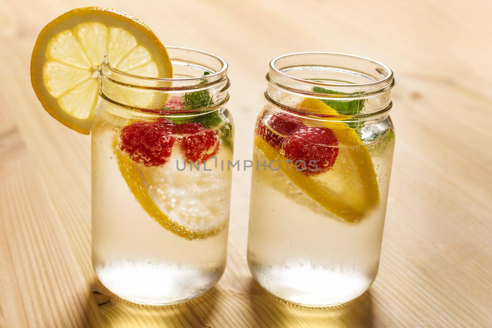 two glass jars with cold water, slices of lemon, mint and red berries, illuminated by sunlight on a wooden table with some pieces of citrus, summer refreshments background, copy space