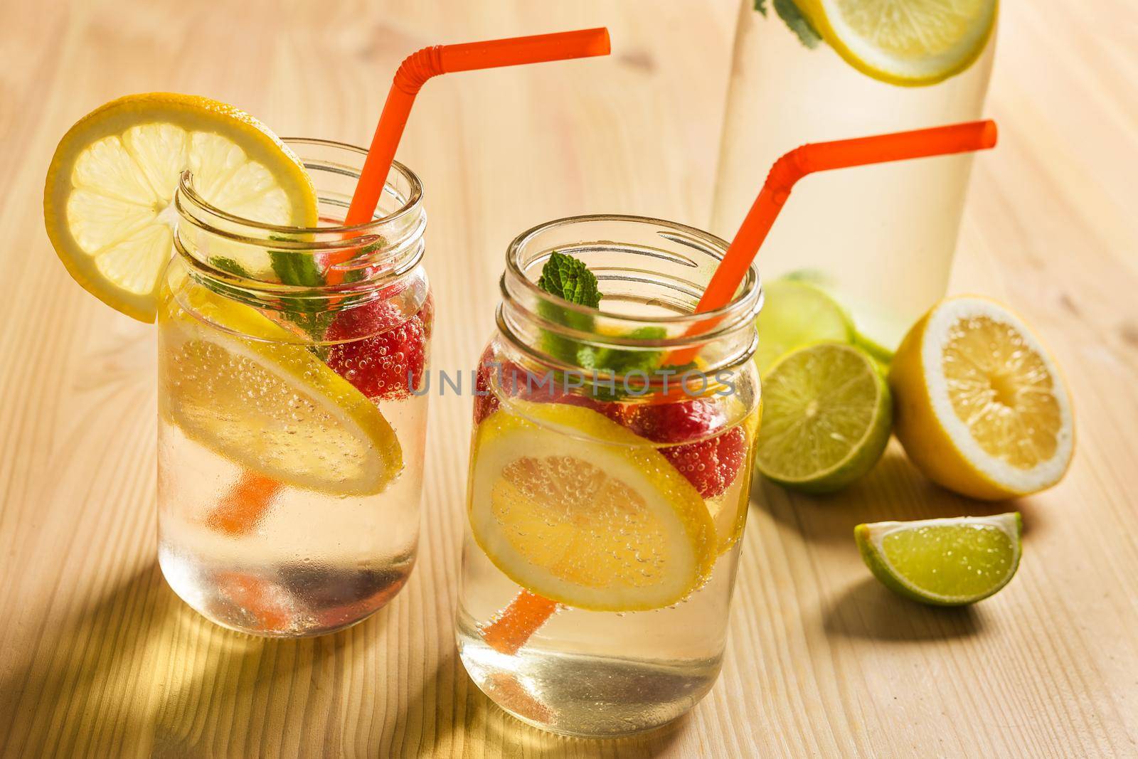 two glass jars and a bottle illuminated by sunlight with refreshing cold lemonade water, lemon slices, red berries, mint leaves and drinking canes on a wooden table. Summer citrus soda background