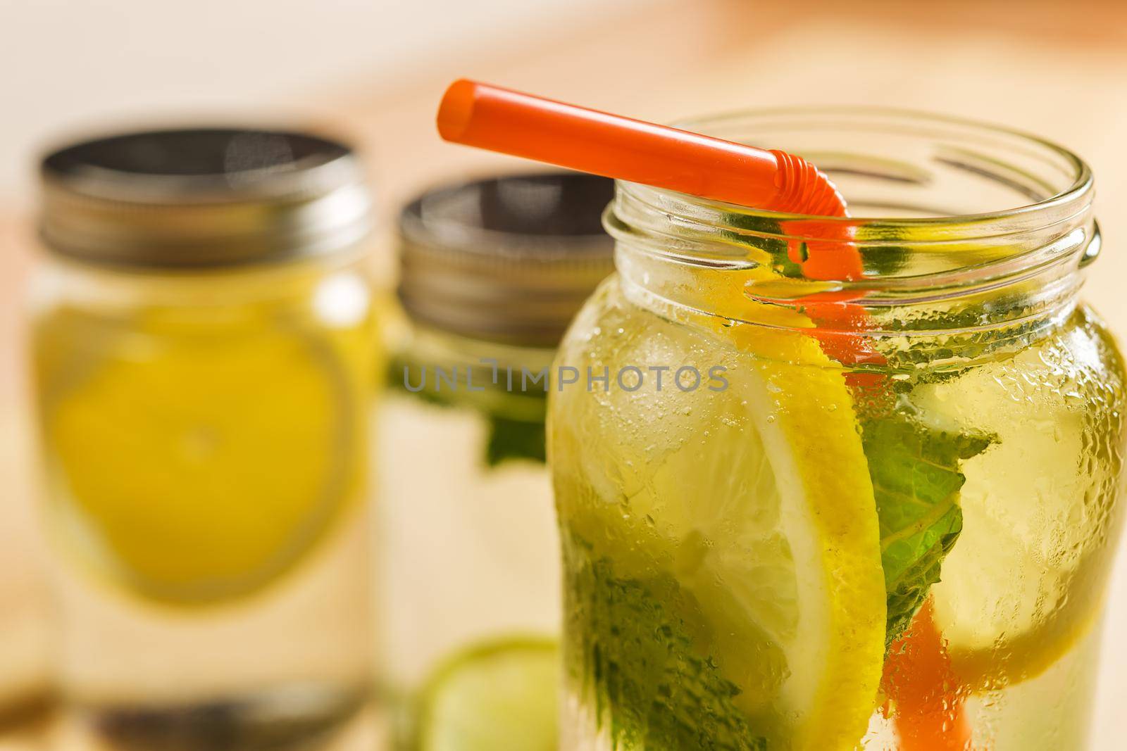 close-up of the top of a glass jar with ice water, lemon, lime, mint leaves and a cane to drink, at the bottom and out of focus are other lemonades