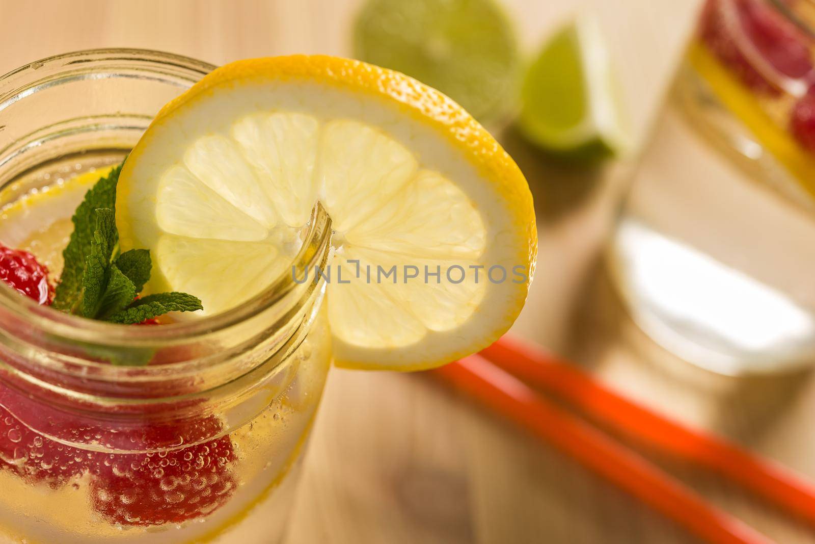 glass jar lit by sunlight with refreshing cold lemonade water, lemon slices, red berries and mint leaves on a unfocused wooden table with drink cane and pieces of citrus. Summer refreshment background