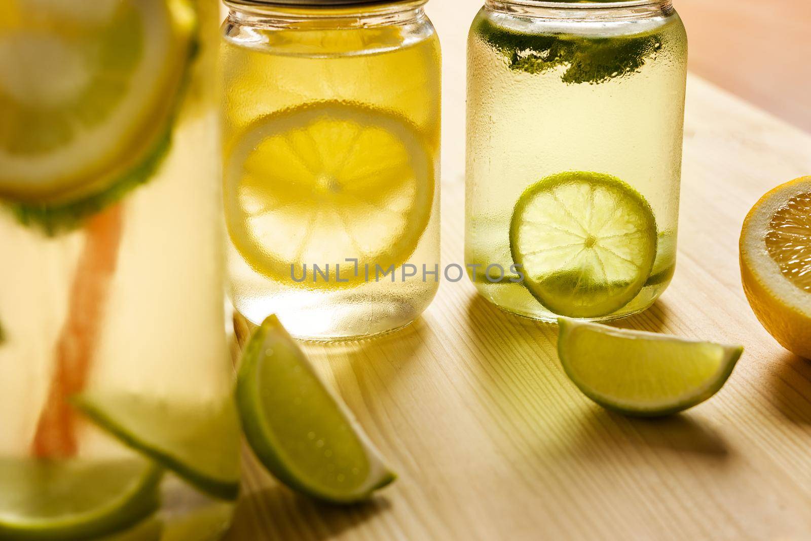 two glass jars with lids illuminated by sunlight with homemade lime and lemon sodas, on the wooden table there are also pieces of fruit and another unfocused lemonade in the foreground