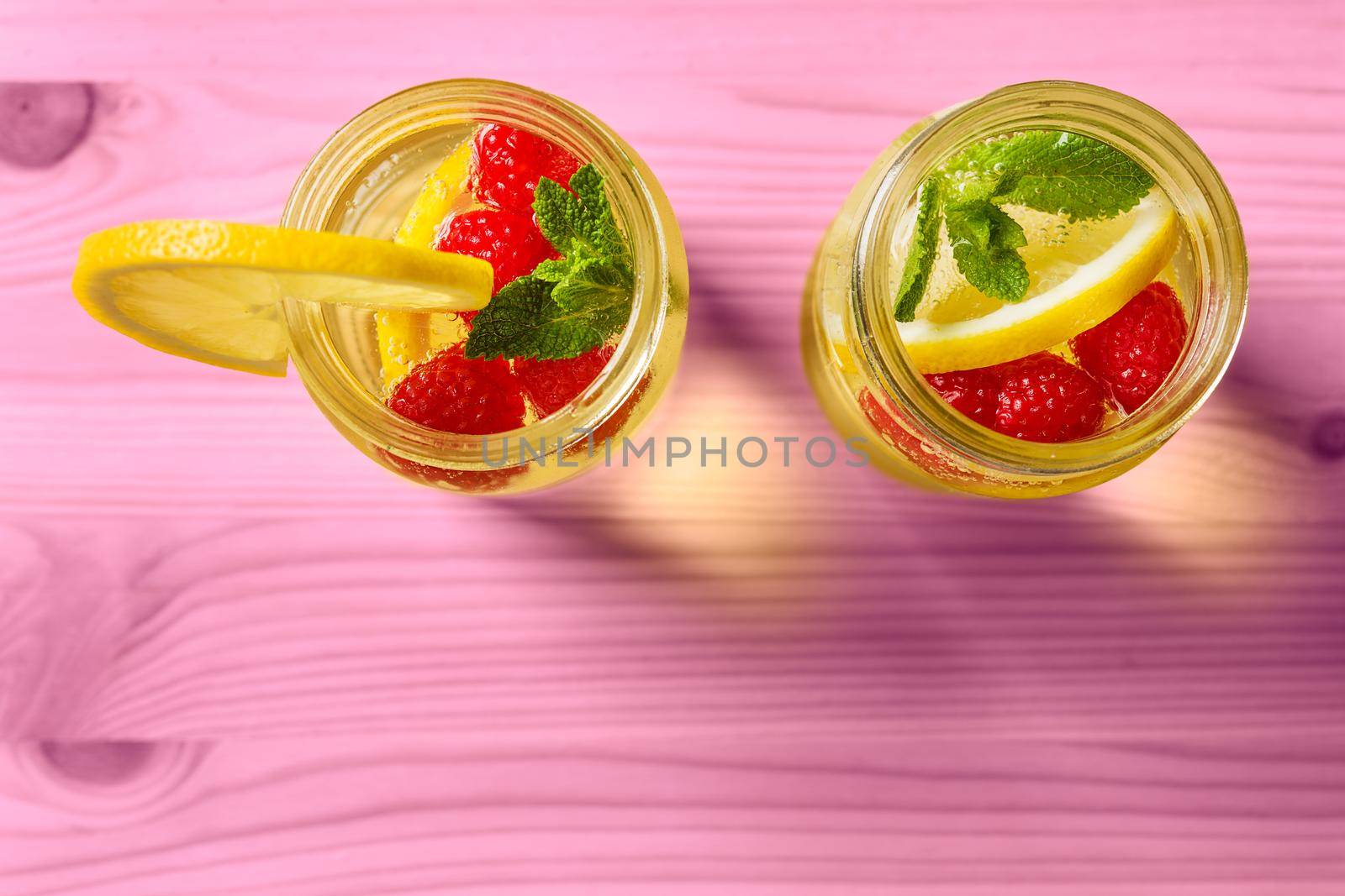 overhead shot of two glass jars with cold water, lemon slices, red berries and mint leaves on a pink wooden table, are illuminated by sunlight. Summer refreshment background with citrus, copy space