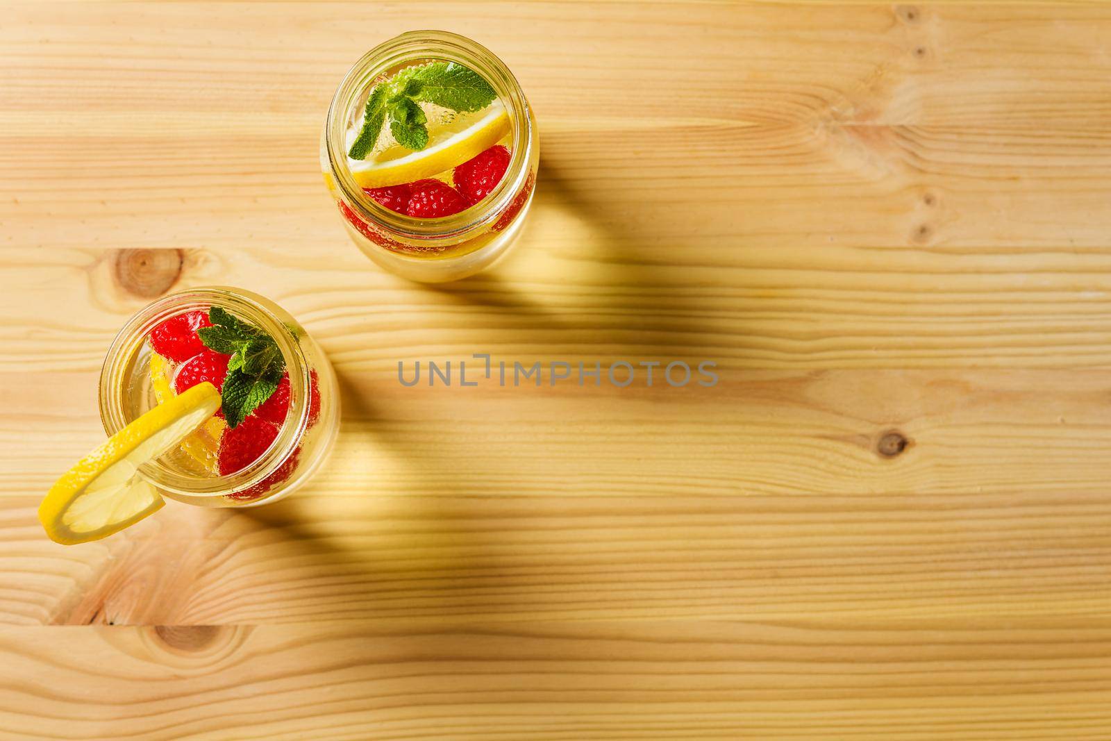 overhead shot of two glass jars with cold water, lemon slices, red berries and mint leaves on one side of a wooden table illuminated by sunlight. Summer refreshment background with citrus, copy space