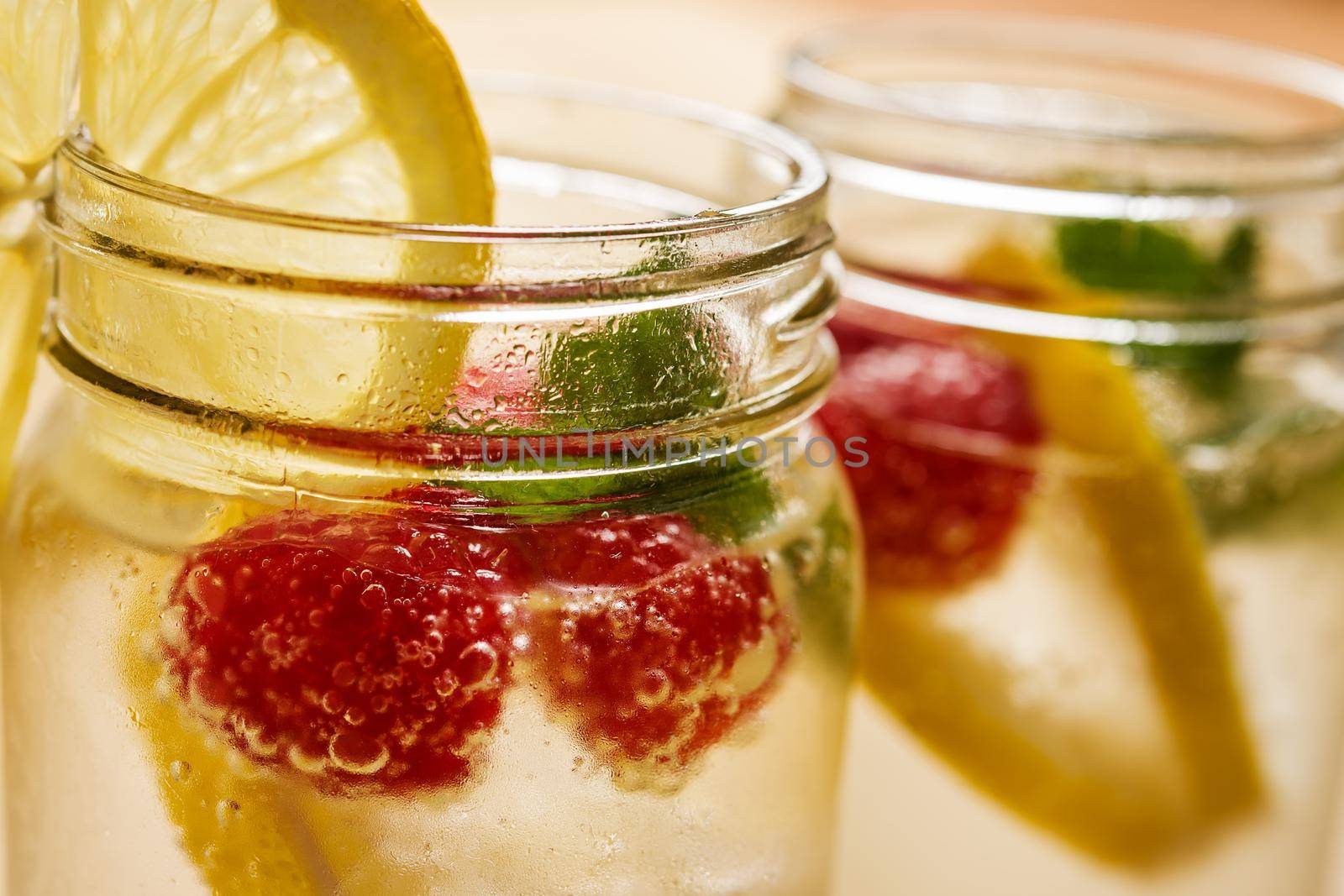 close-up of a glass jar with cold water, slices of lemon, mint and red berries, illuminated by sunlight on a wooden table, in the background it looks out of focus another glass equal