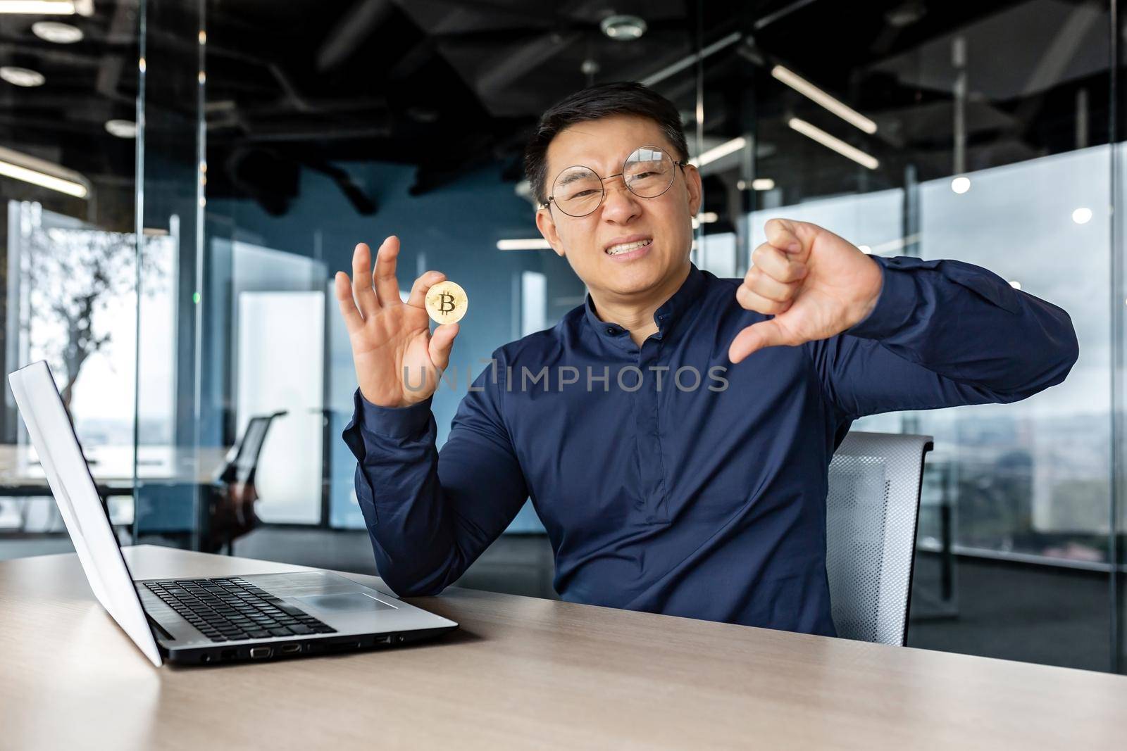 Dissatisfied asian businessman holding finger down, bad losing gesture, holding virtual currency bitcoin money, man looking disappointed at camera, working inside modern office building.