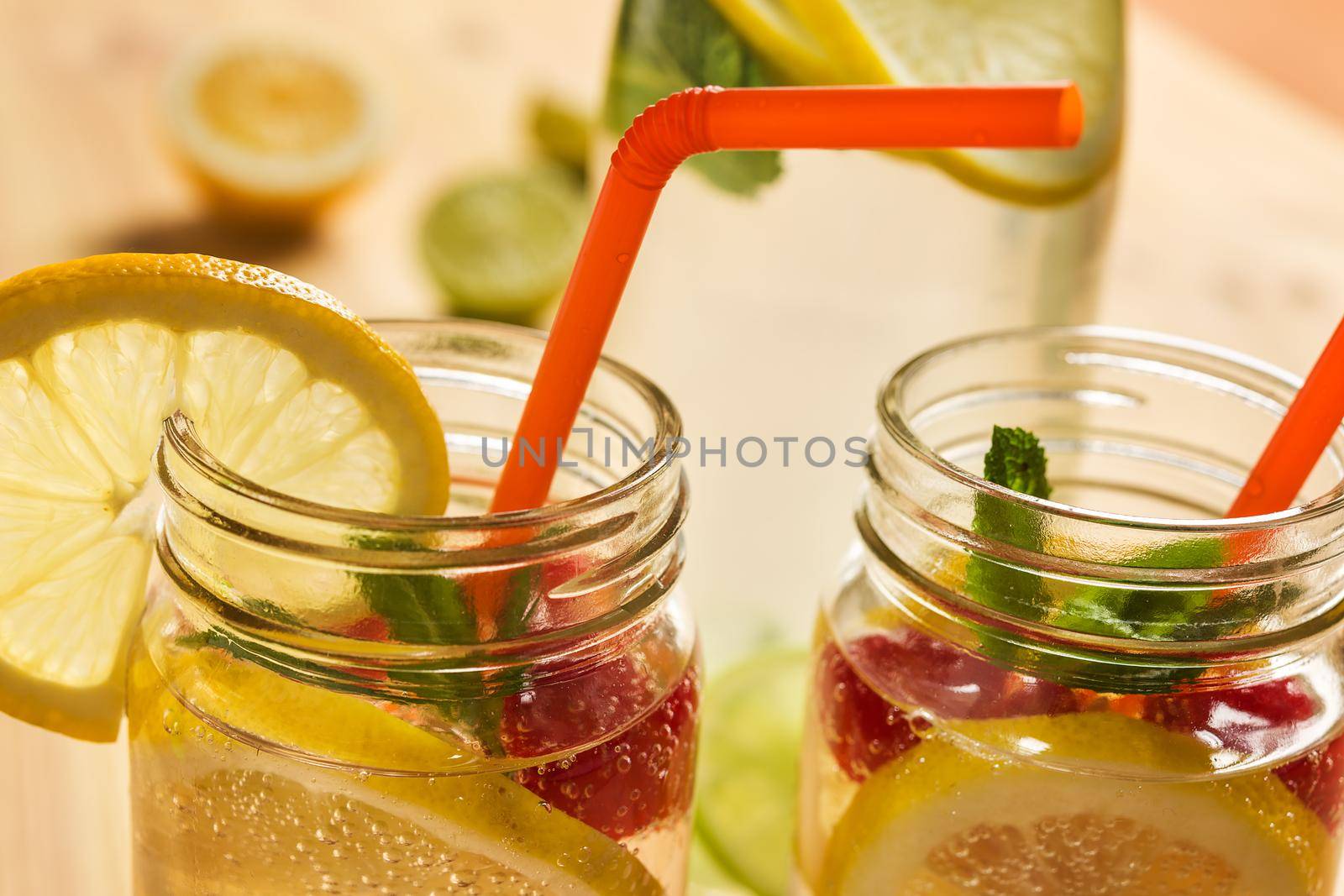 close-up of two glass jars illuminated by sunlight with refreshing cold lemonade water, lemon slices, red berries, mint leaves and drink canes. Summer citrus soda background