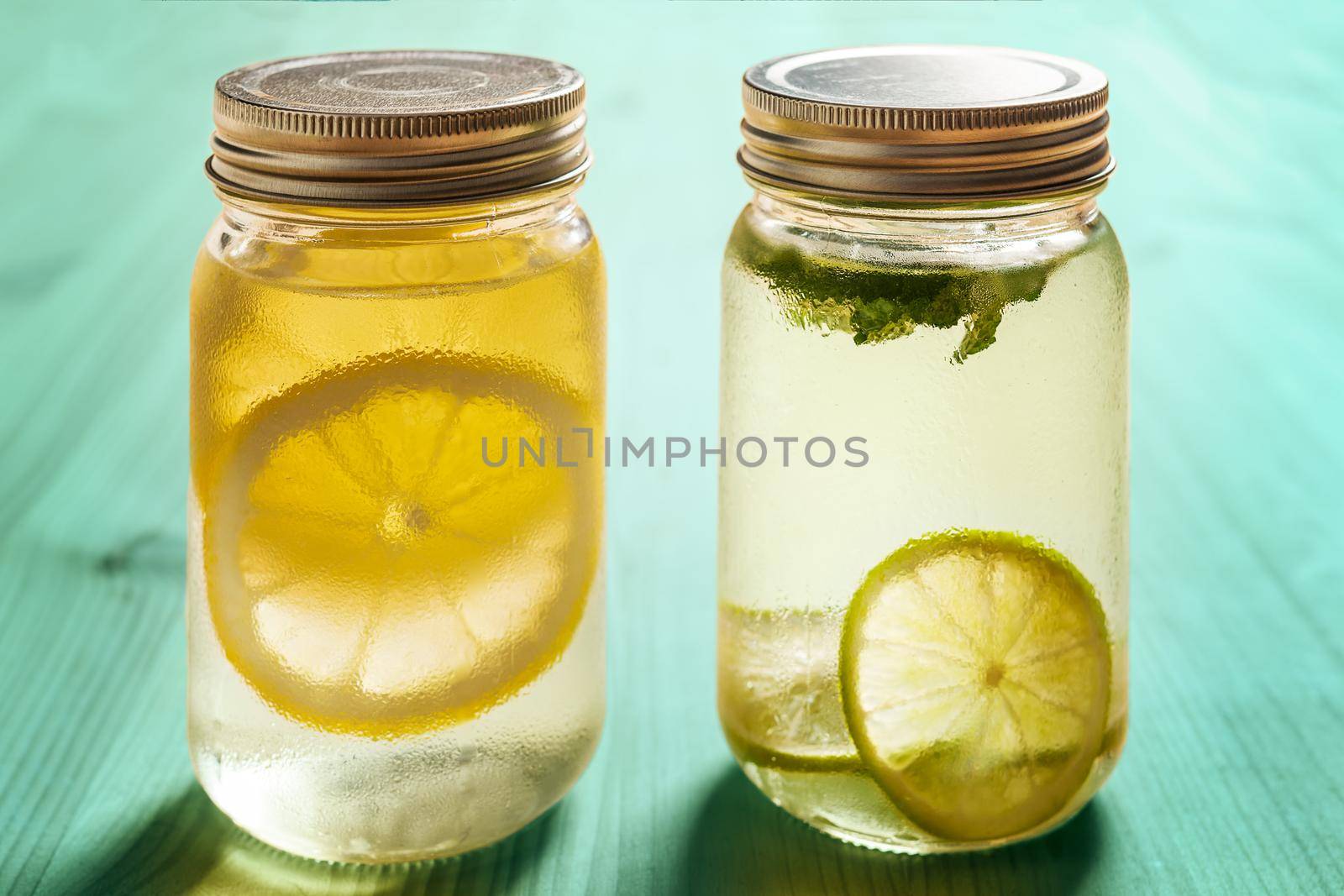 two glass jars with lids on a turquoise wooden table illuminated by sunlight, one has cold water with lemon and the other with lime and mint