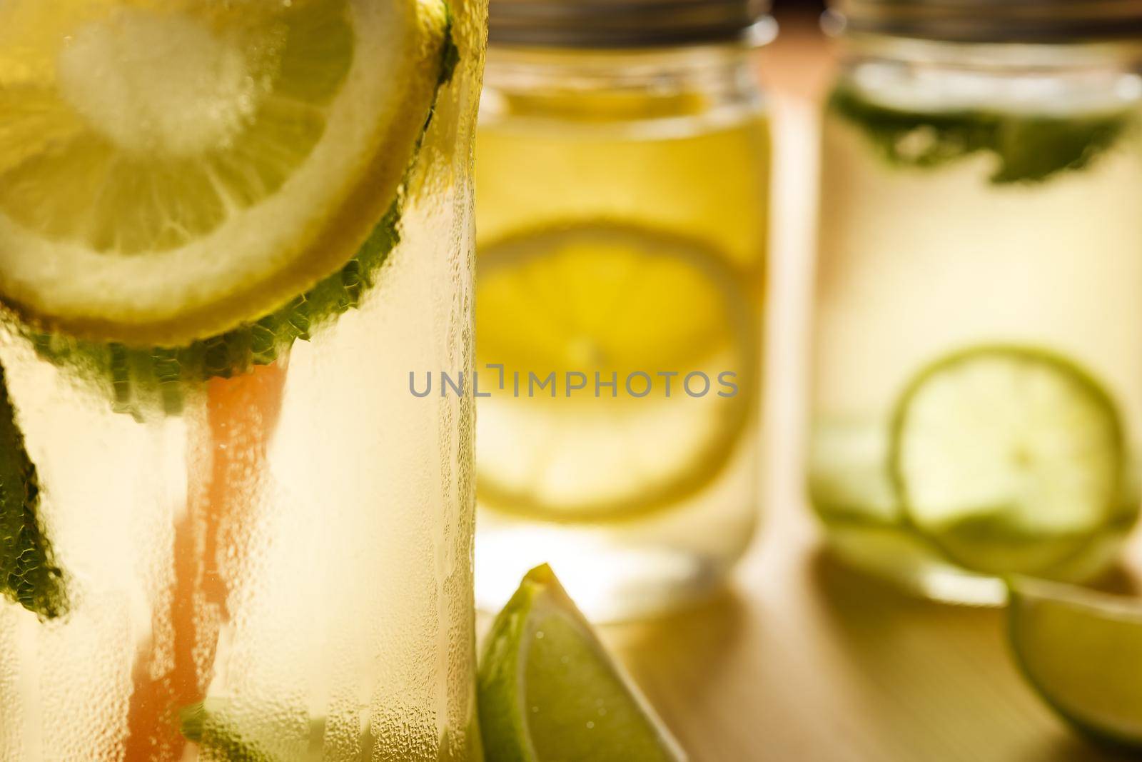 close-up of an iced lemonade with an unfocused background of two glass jars with lid on a wooden table, the jars contains homemade lime and lemon sodas