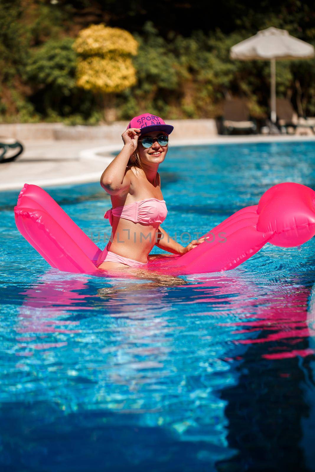 Sexy woman in sunglasses with a smile on her face in a swimsuit lies on a pink inflatable mattress in the pool. Relax by the pool on a hot summer sunny day. Vacation concept by Dmitrytph