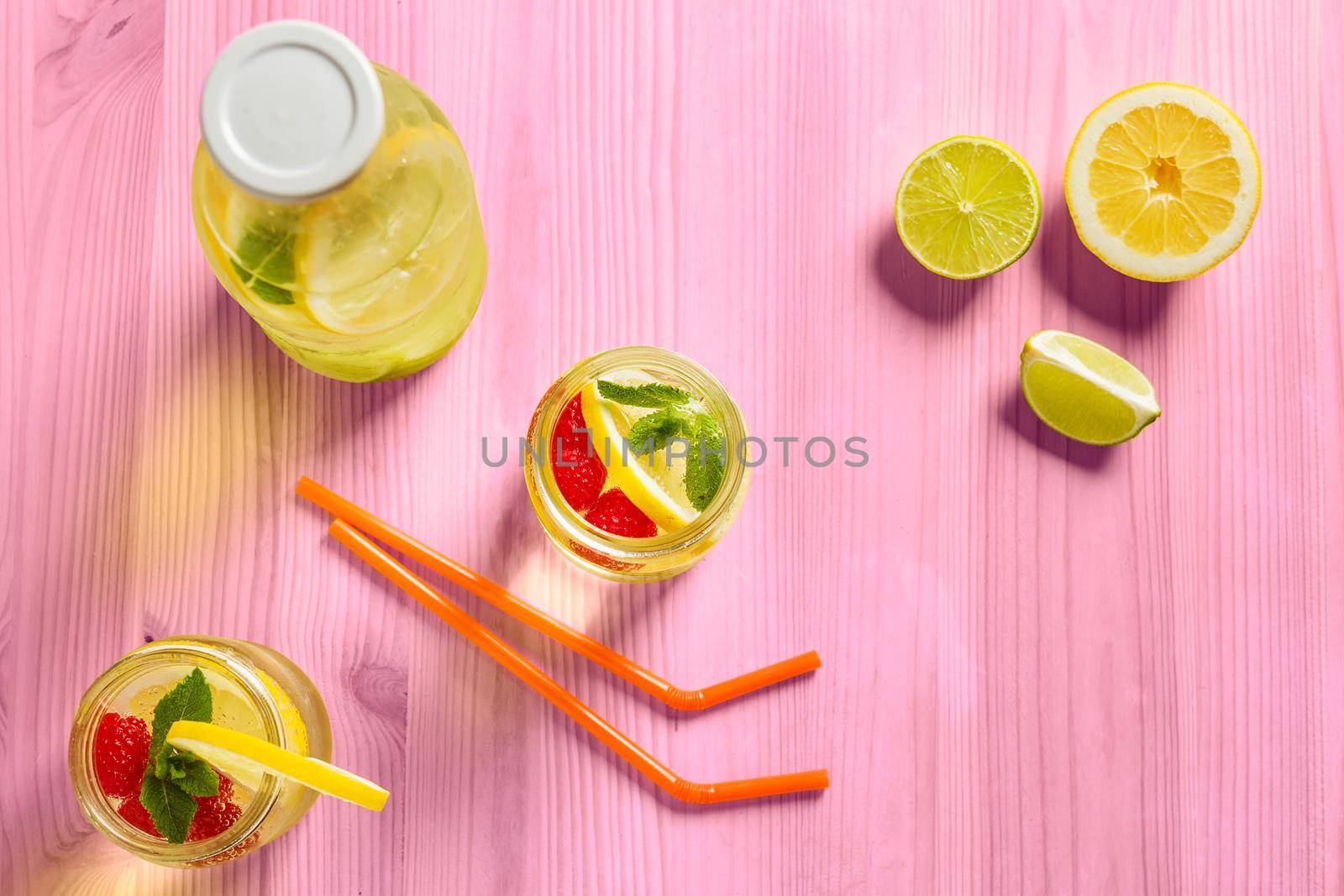 top view, two glass jars and a bottle with refreshing cold lemonade water, lemon, red berries and mint on a pink wooden table with citrus lit by sunlight, Summer refreshment background Copy space