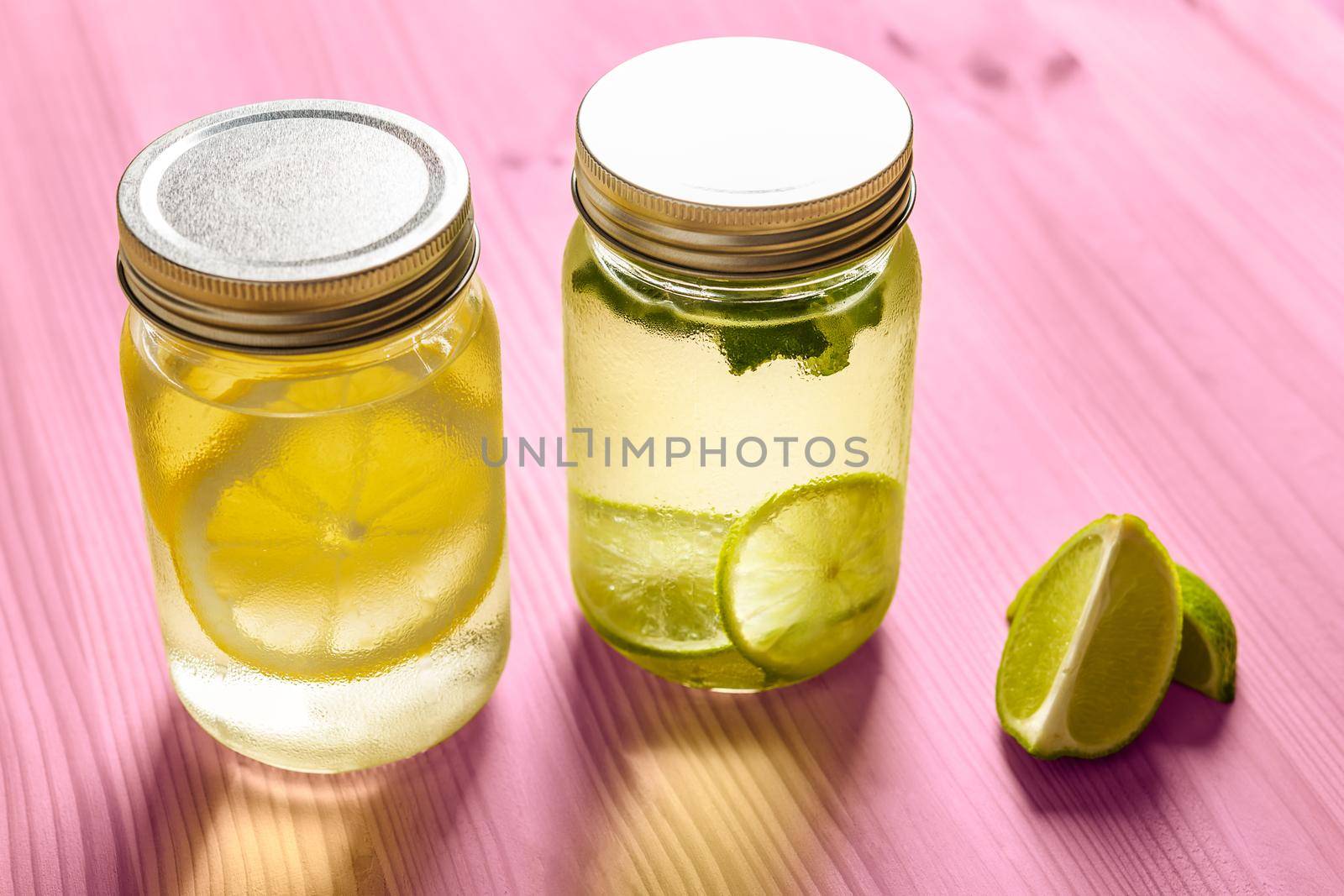 two glass jars with lids full of water with slices of lime and lemon, are on a pink wooden table and illuminated by sunlight, there are also two slices of lime on the table