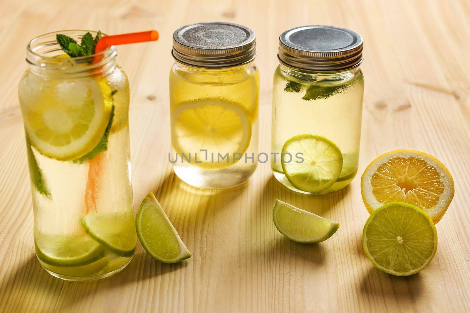 two glass jars with lids illuminated by sunlight with homemade lime and lemon sodas, on the wooden table there are also pieces of fruit and another lemonade with mint leaves and a cane