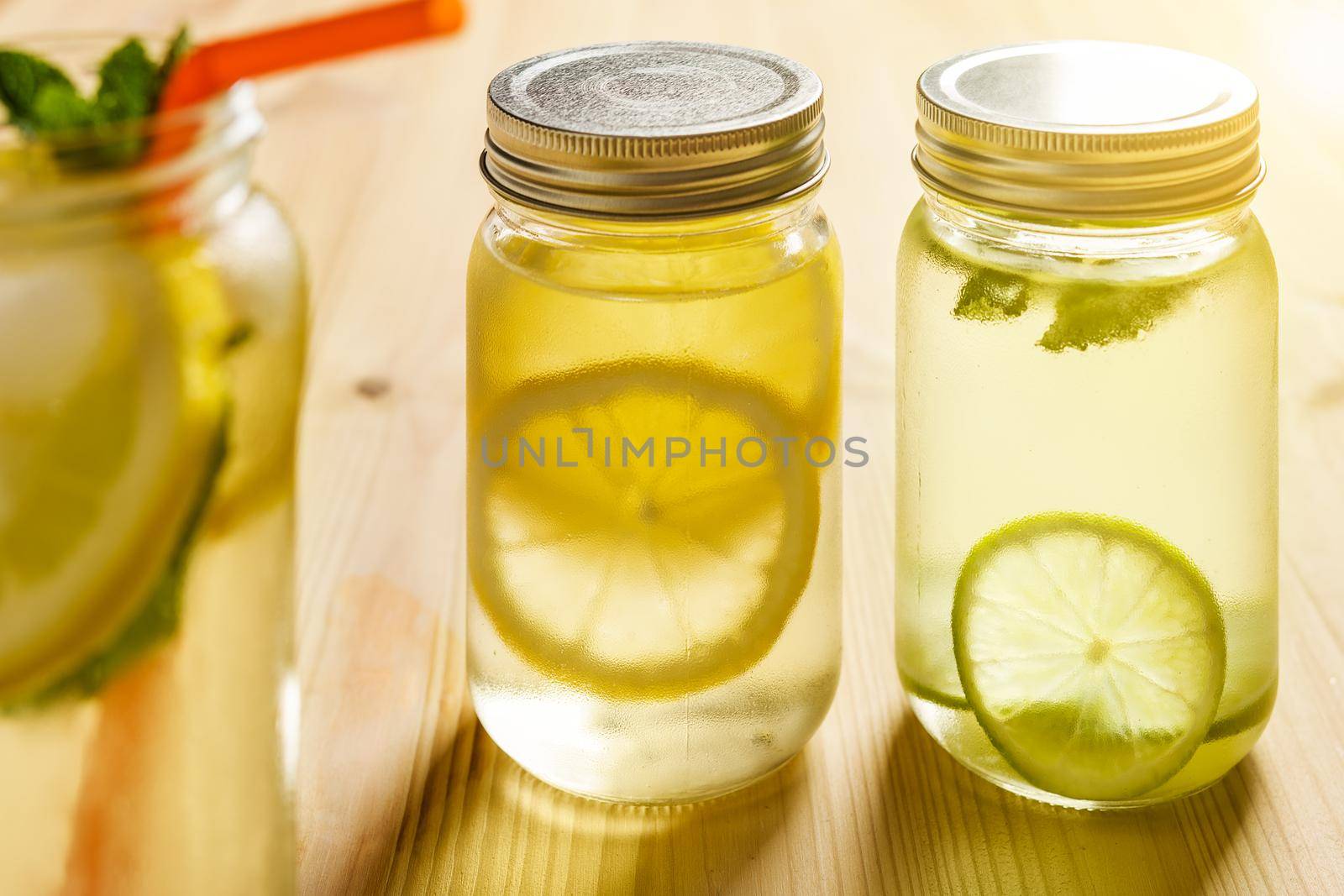 two glass jars with lids illuminated by sunlight with homemade lime and lemon sodas, in the foreground there is another lemonade out of focus