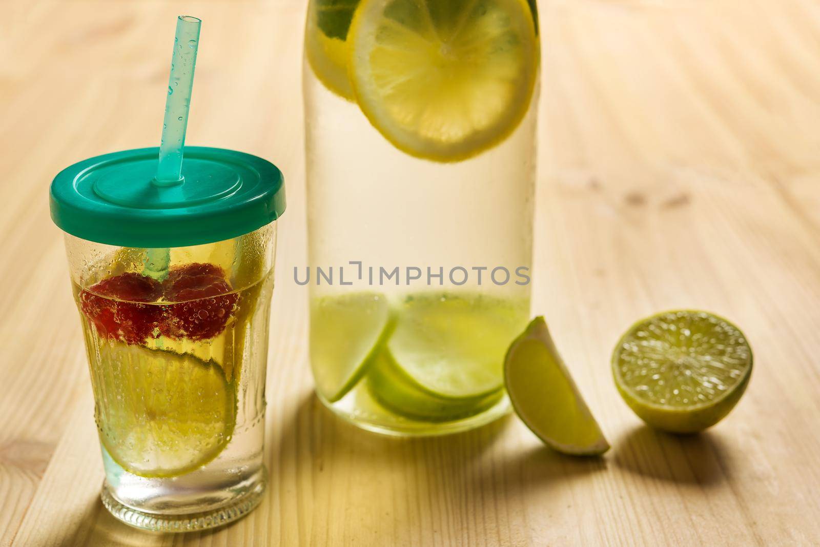 bottle and glass with lid and cane with cold water and slices of lime, lemon and red berries, illuminated by sunlight on a wooden table with some pieces of citrus, summer refreshments background