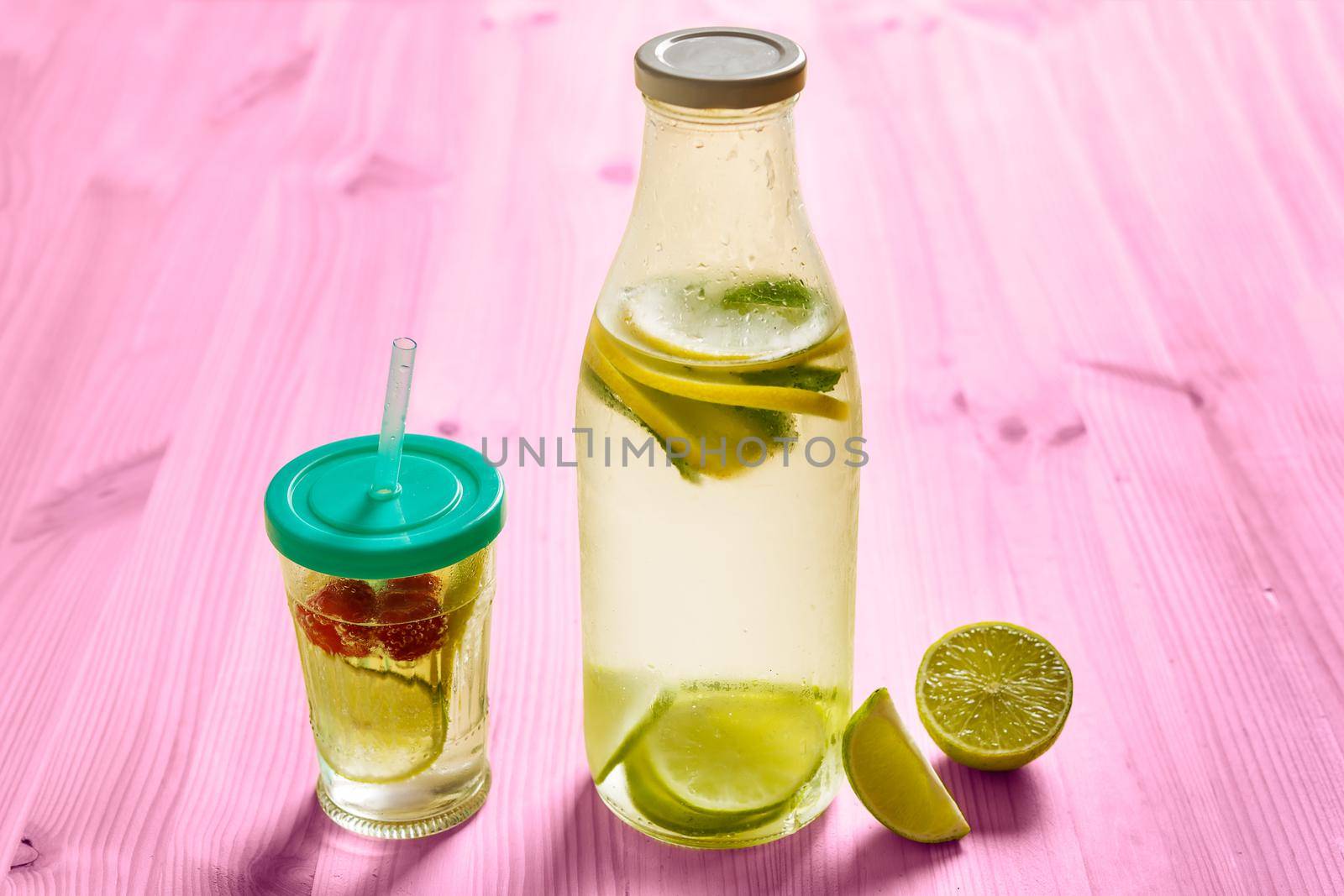 glass bottle and glass with lid and cane, with cold water and slices of lemon, lime, berries and mint, are illuminated by sunlight on a pink wooden table with some pieces of citrus