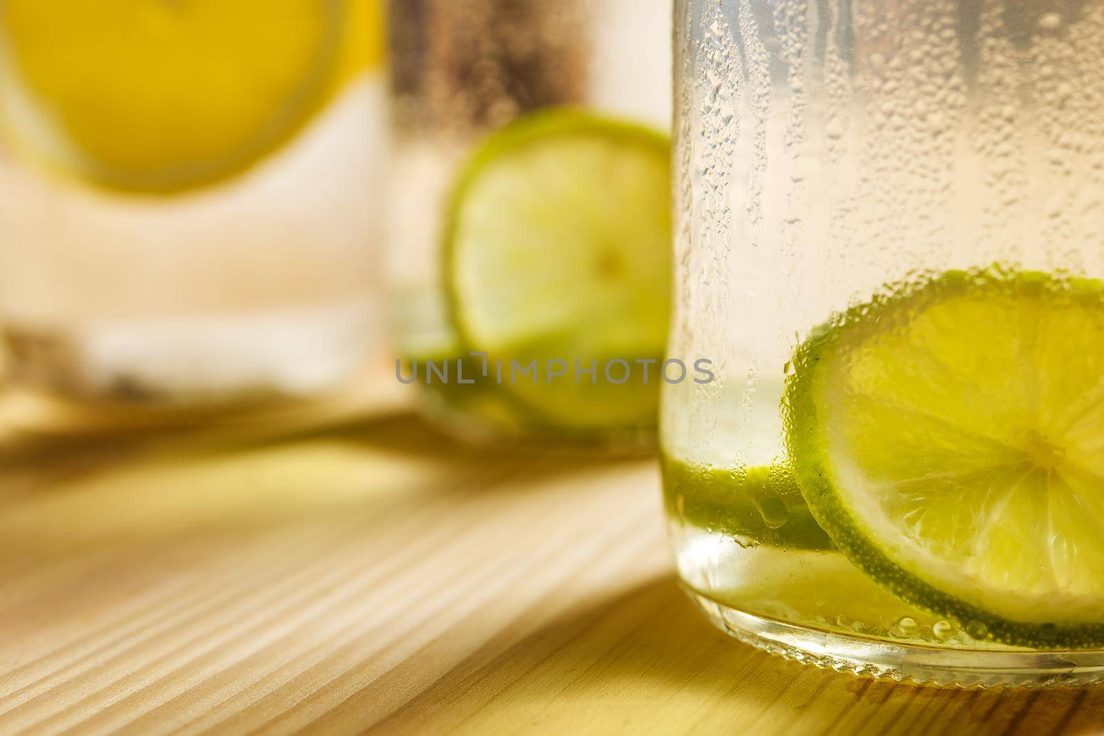 close-up of the bottom of a glass jar with ice water, lemon, lime and mint leaves on a wooden table and illuminated by sunlight, in the background and out of focus are other lemonades