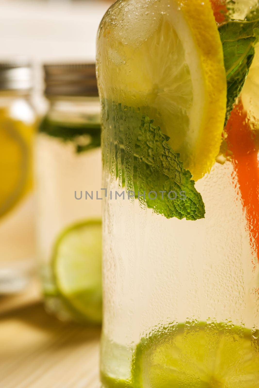 vertical close-up of a glass jar with ice water, lemon, lime and mint leaves, it is on a wooden table and illuminated by sunlight, in the background and out of focus are other lemonades
