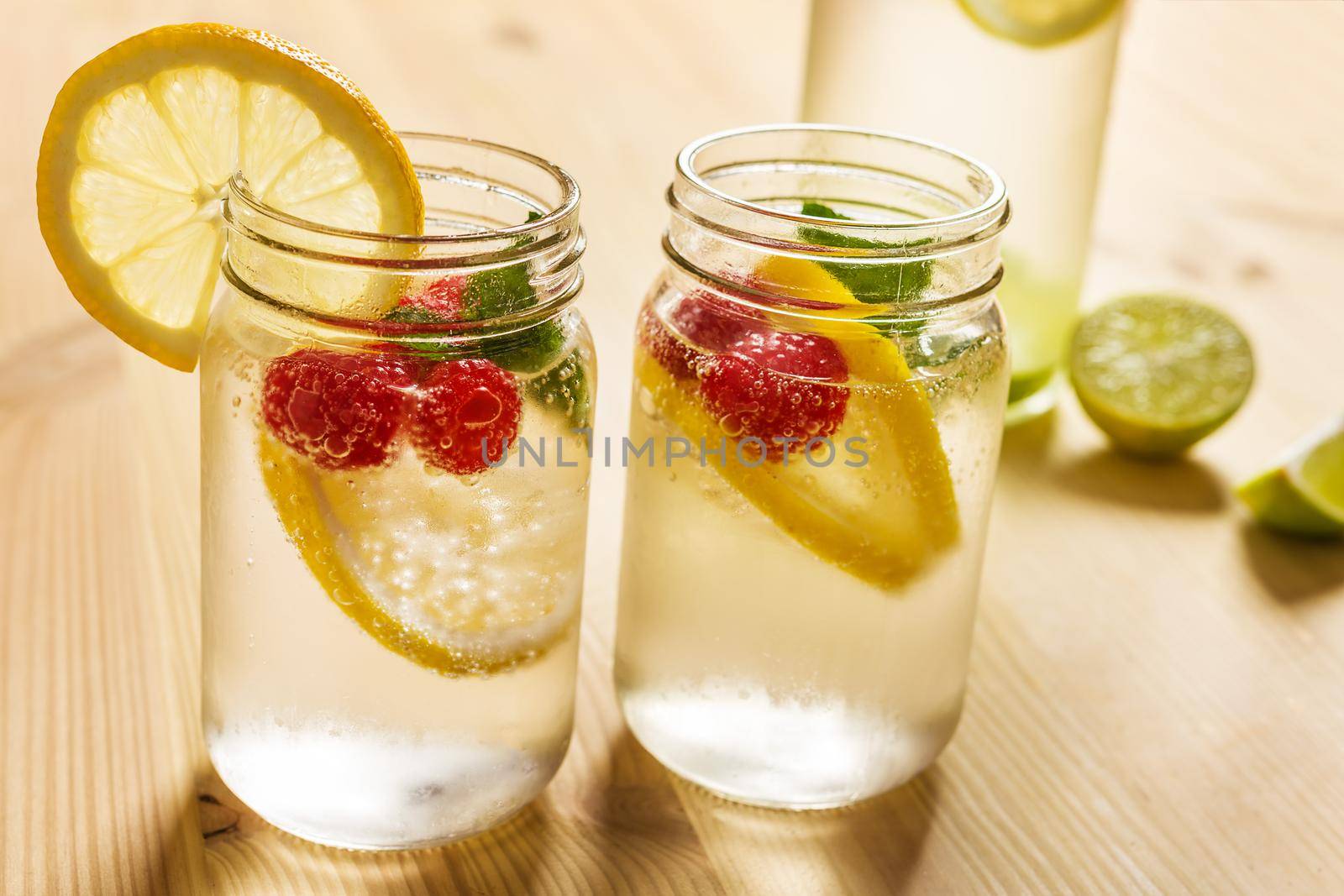 two glass jars with cold water, slices of lime, lemon, mint and red berries illuminated by sunlight on a wooden table with a bottle and pieces of citrus, summer refreshments background, copy space