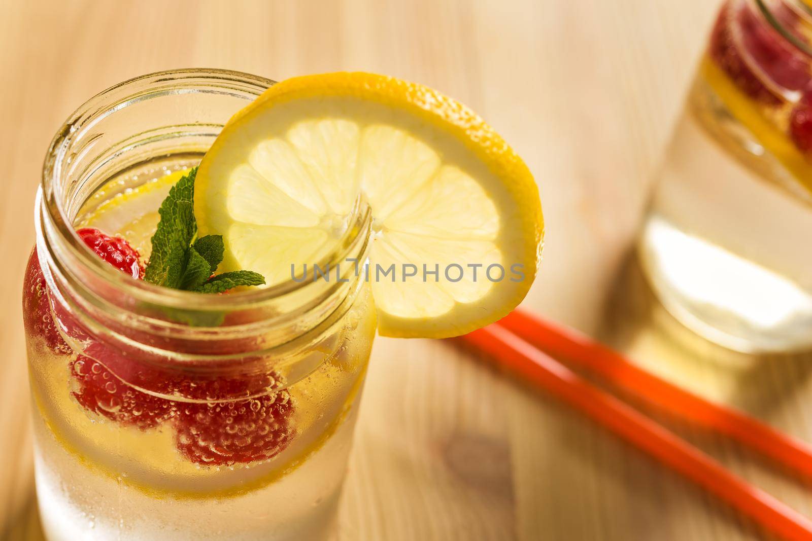 glass jar illuminated by sunlight with refreshing cold lemonade water, lemon slices, red berries and mint leaves on a wooden table with drink canes. Summer citrus soda background. copy space