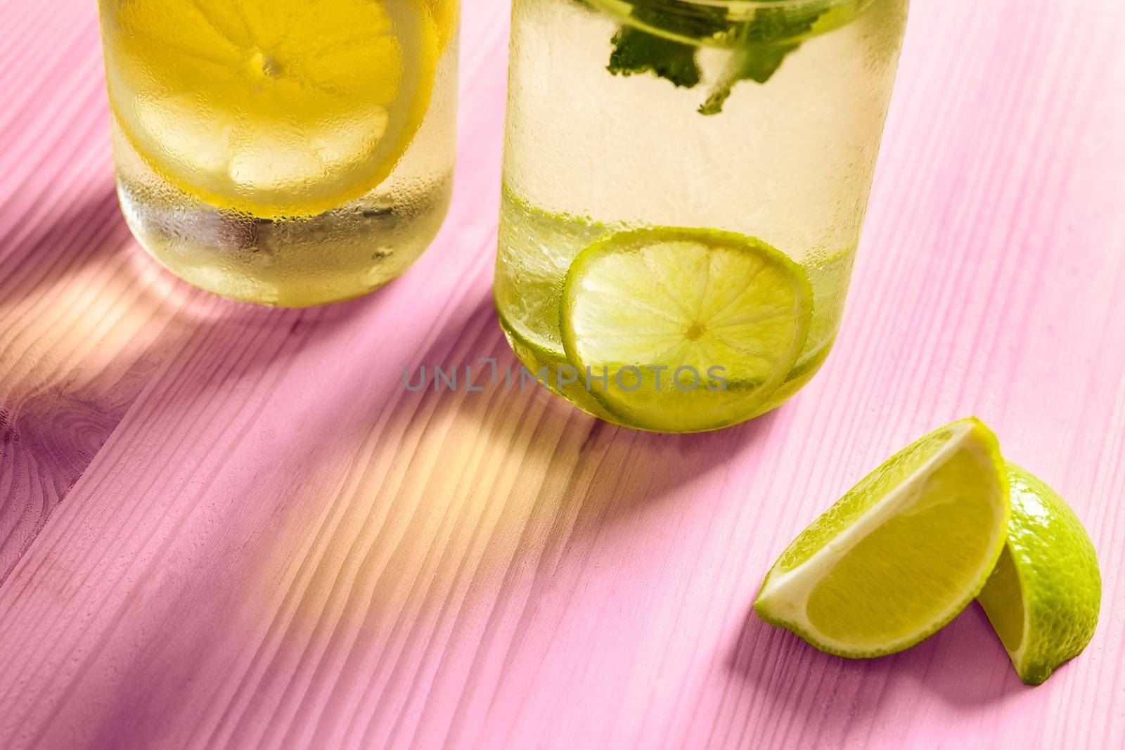 cold summer refreshments with slices of citrus and mint leaves, are on a pink wooden table and illuminated by sunlight, there are also lemon wedges and copy space