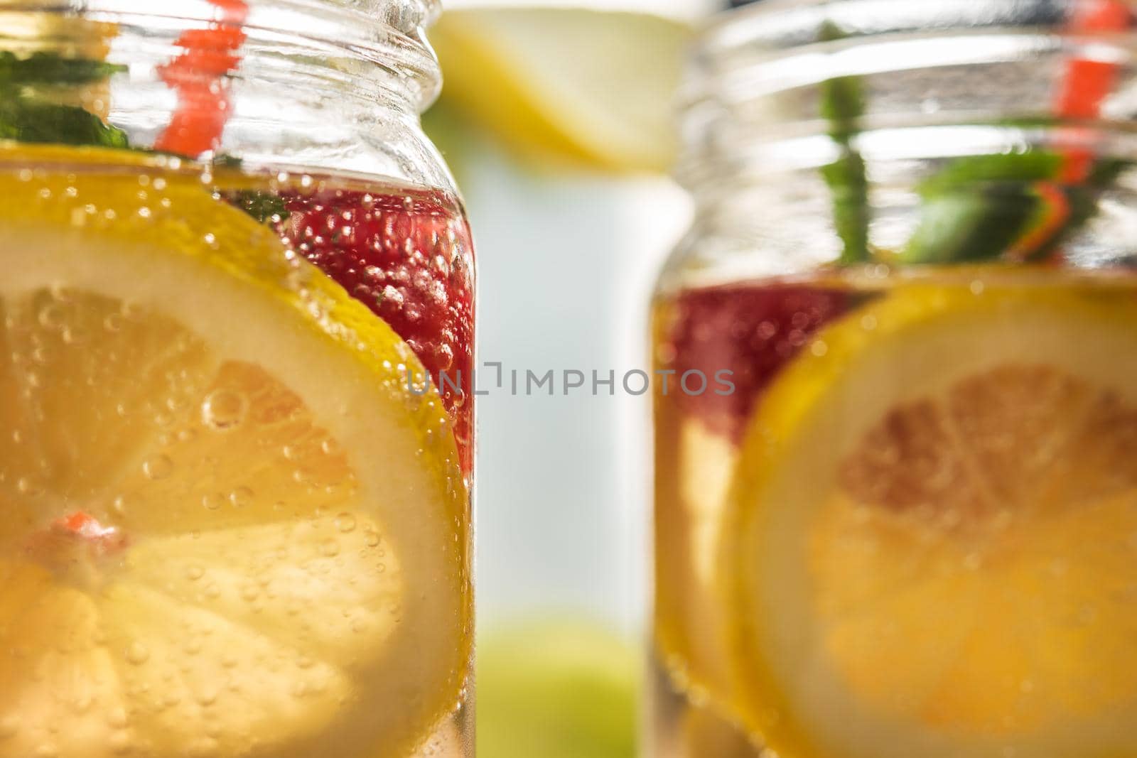 close-up of two glass jars illuminated by sunlight with refreshing cold lemonade water, lemon slices, red berries, mint leaves and drinking canes. Summer citrus soda background