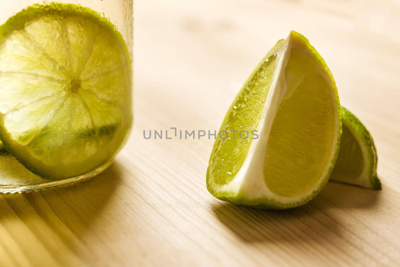 horizontal photo of some slices of lime next to a glass jar with lemonade, this one is illuminated by sunlight and on a wooden table