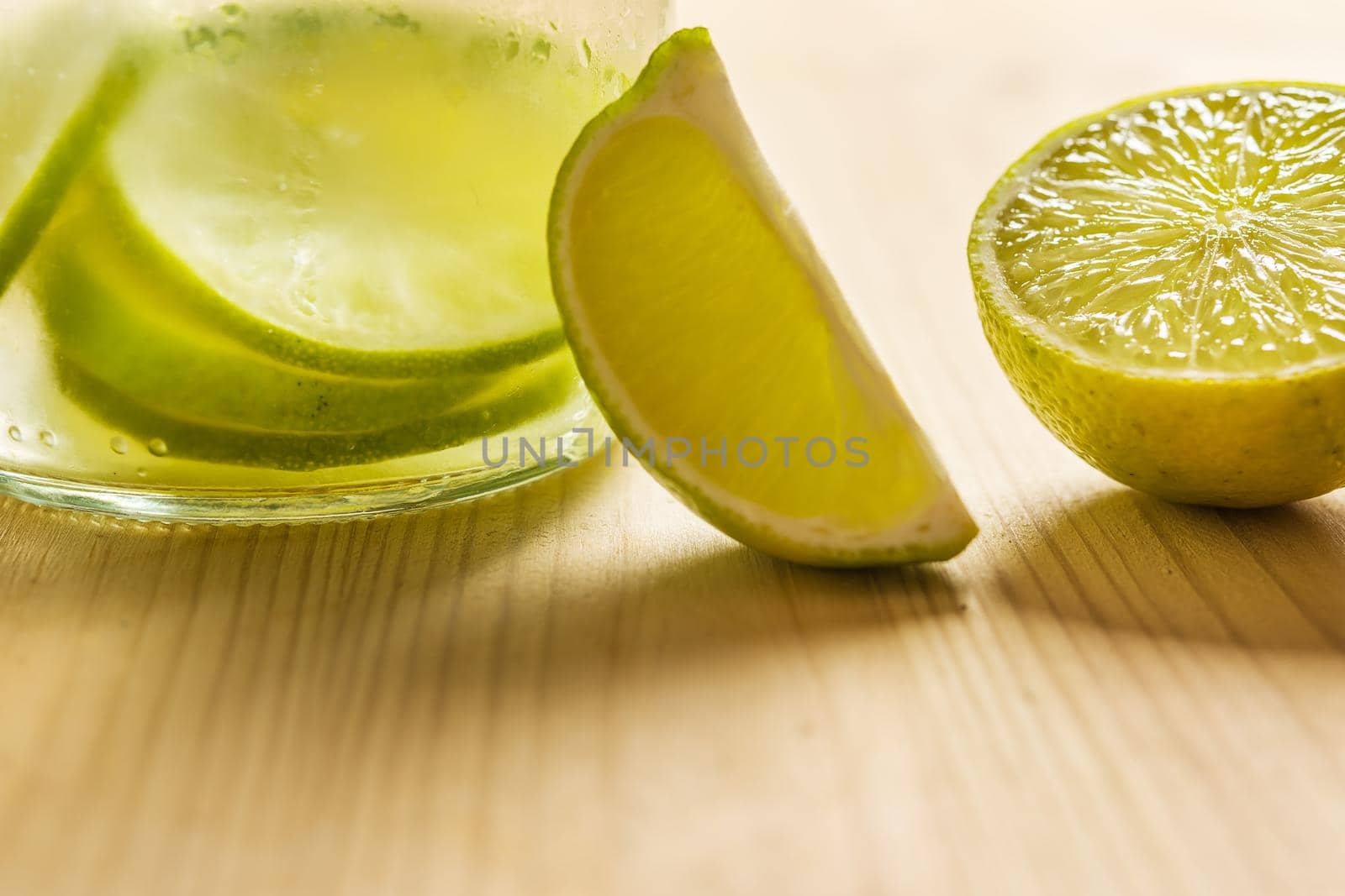 Glass bottle with cold water and slices of lime and lemon, illuminated by sunlight on a wooden table with some pieces of citrus, citrus summer refreshments background, copy space