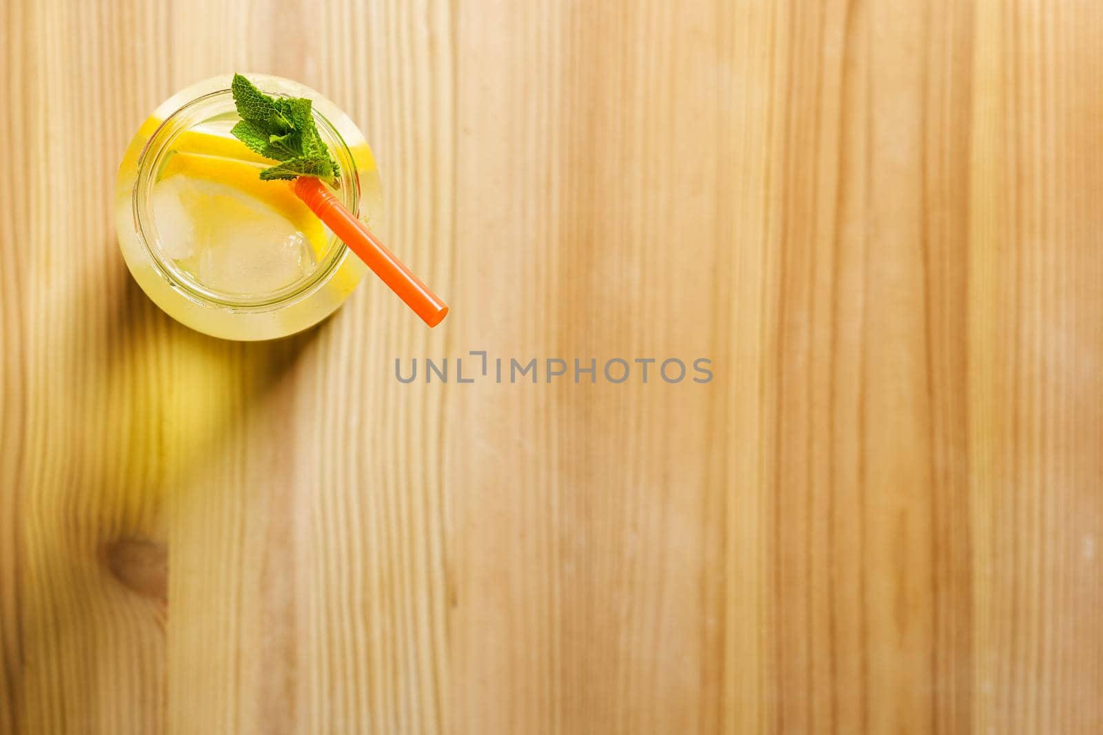 Flat lay of a glass jug with lemonade on a wooden table, the glass contains water, lemon, mint, ice and a straw.