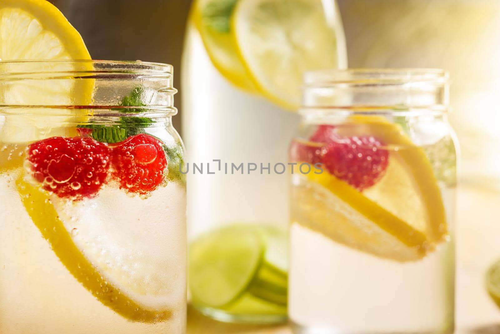 Two glass jars with refreshing cold water, lemon slices, red berries, lime and mint and a bottle with citrus pieces illuminated by sunlight. Summer refreshment background. copy space