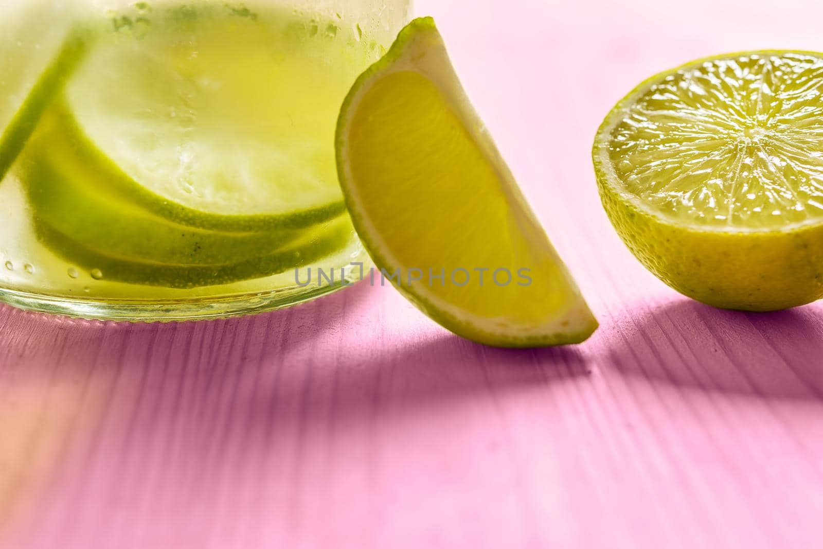 Glass bottle with cold water and slices of lime and lemon, illuminated by sunlight on a pink wooden table with some pieces of citrus, citrus summer refreshments background, copy space