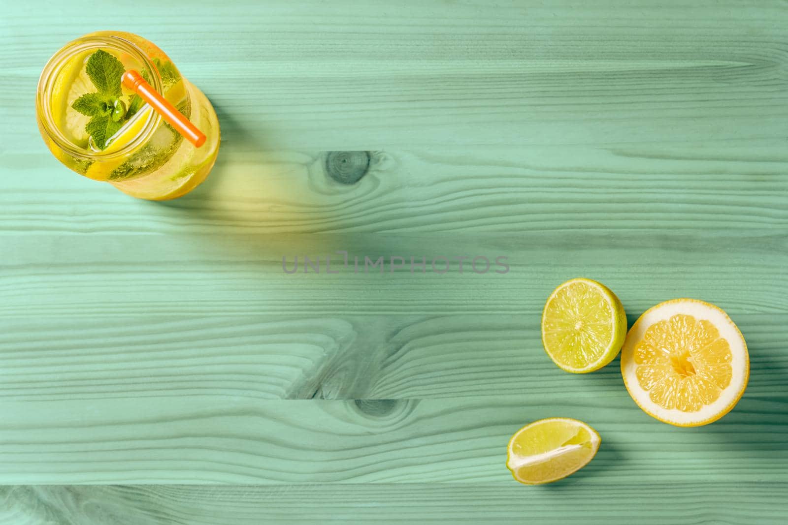 flat lay of homemade lemonade with mint and a cane to drink in a glass jar lit by sunlight, it's on a turquoise wooden table, there are also a few pieces of lime and lemon and copy space