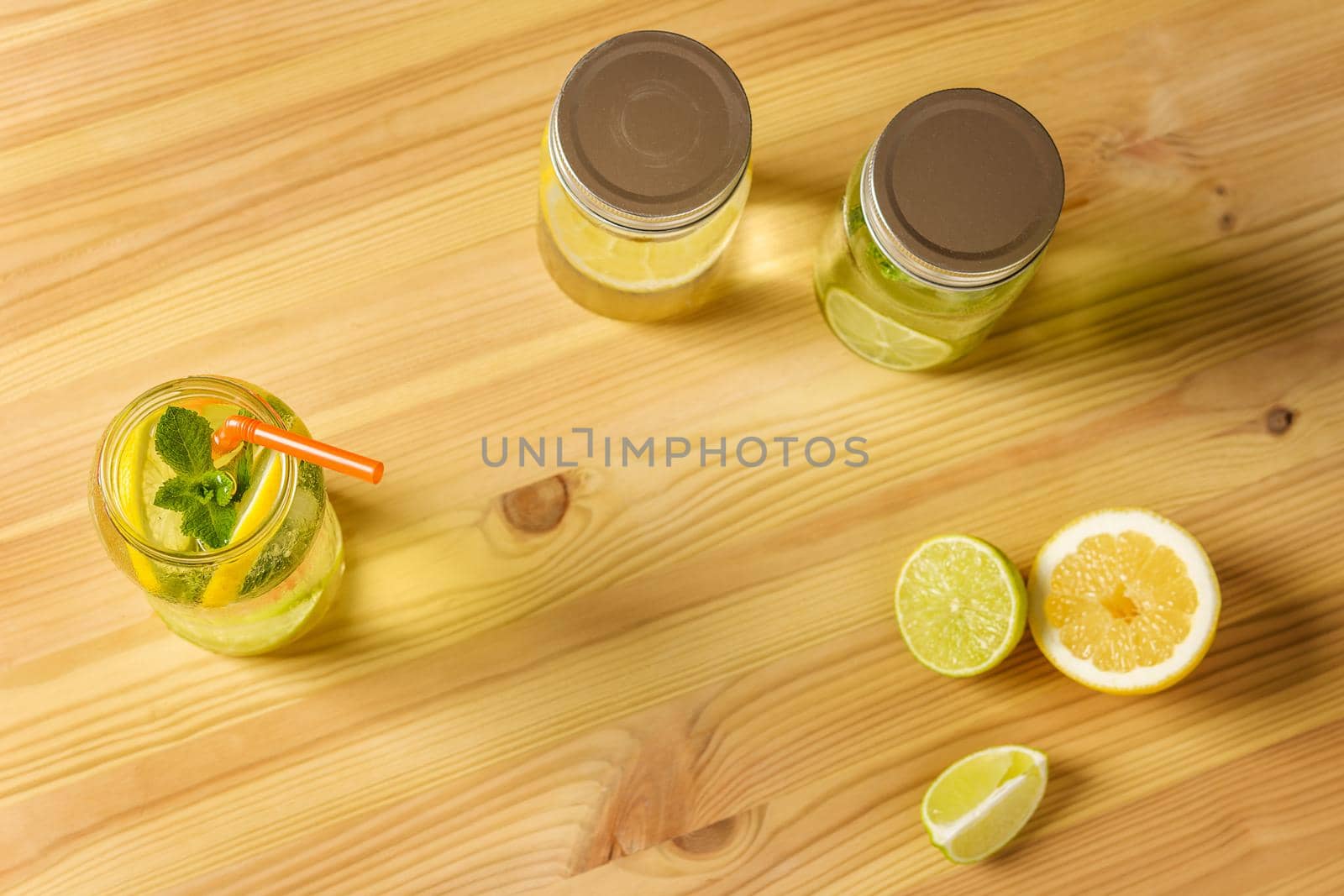 Flat Lay of lemonade with mint and a cane to drink in a glass jar illuminated by sunlight, is on a wooden table, there are also two other jars with lid, some pieces of lemon and lime and copy space