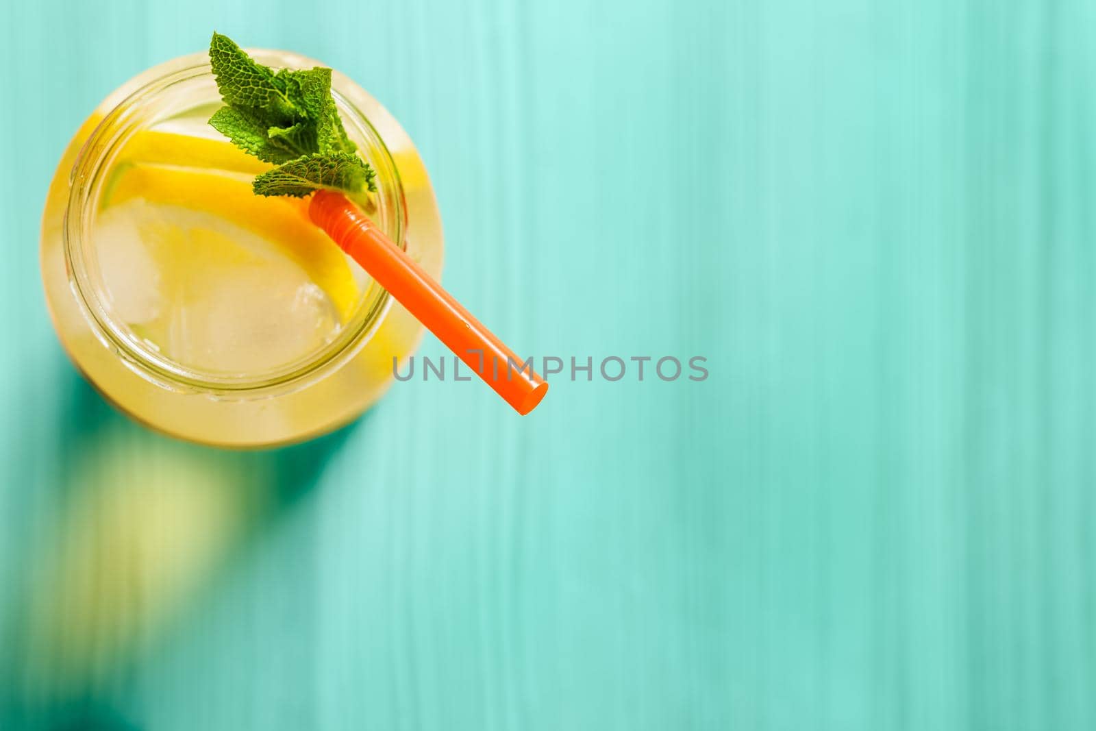 Flat lay of a glass jug with lemonade on a turquoise wooden table, the glass contains water, lemon, mint, ice and a straw.