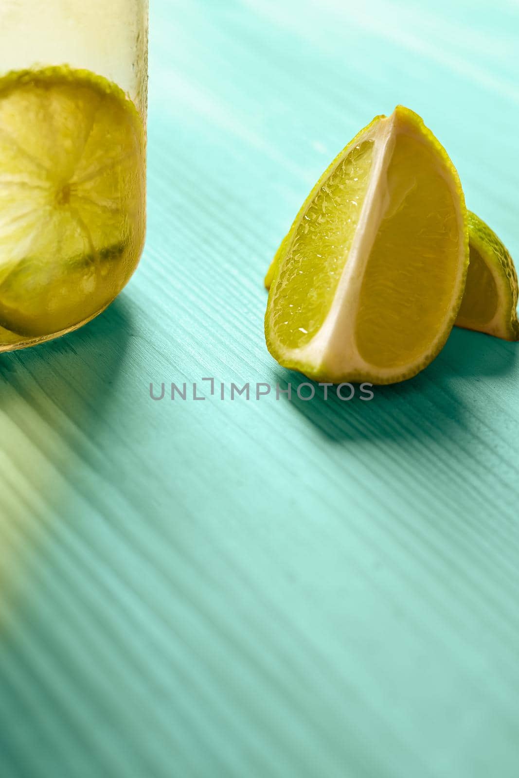 vertical photo with copy space of some slices of lime next to a glass jar with lemonade, this one is illuminated by sunlight and on a turquoise wooden table