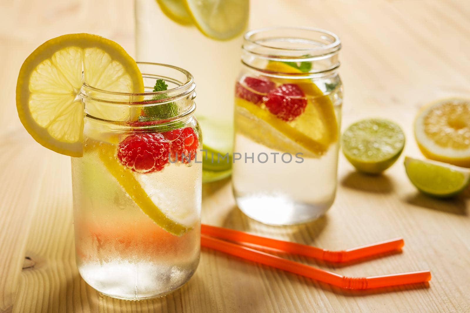 a bottle and two glass jars with cold water, slices of lemon, red berries and mint on a wooden table with canes and citrus pieces illuminated by sunlight. Summer refreshment background with citrus