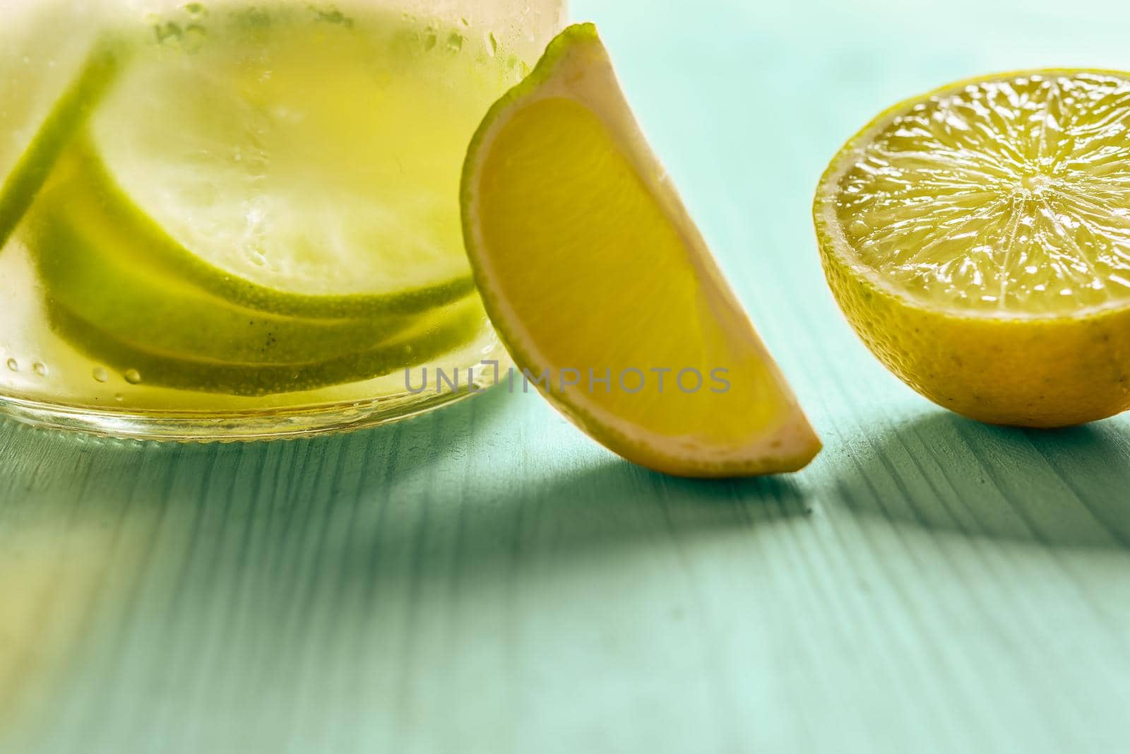 Glass bottle with cold water and slices of lime and lemon, illuminated by sunlight on a green wooden table with some pieces of citrus, citrus summer refreshments background, copy space