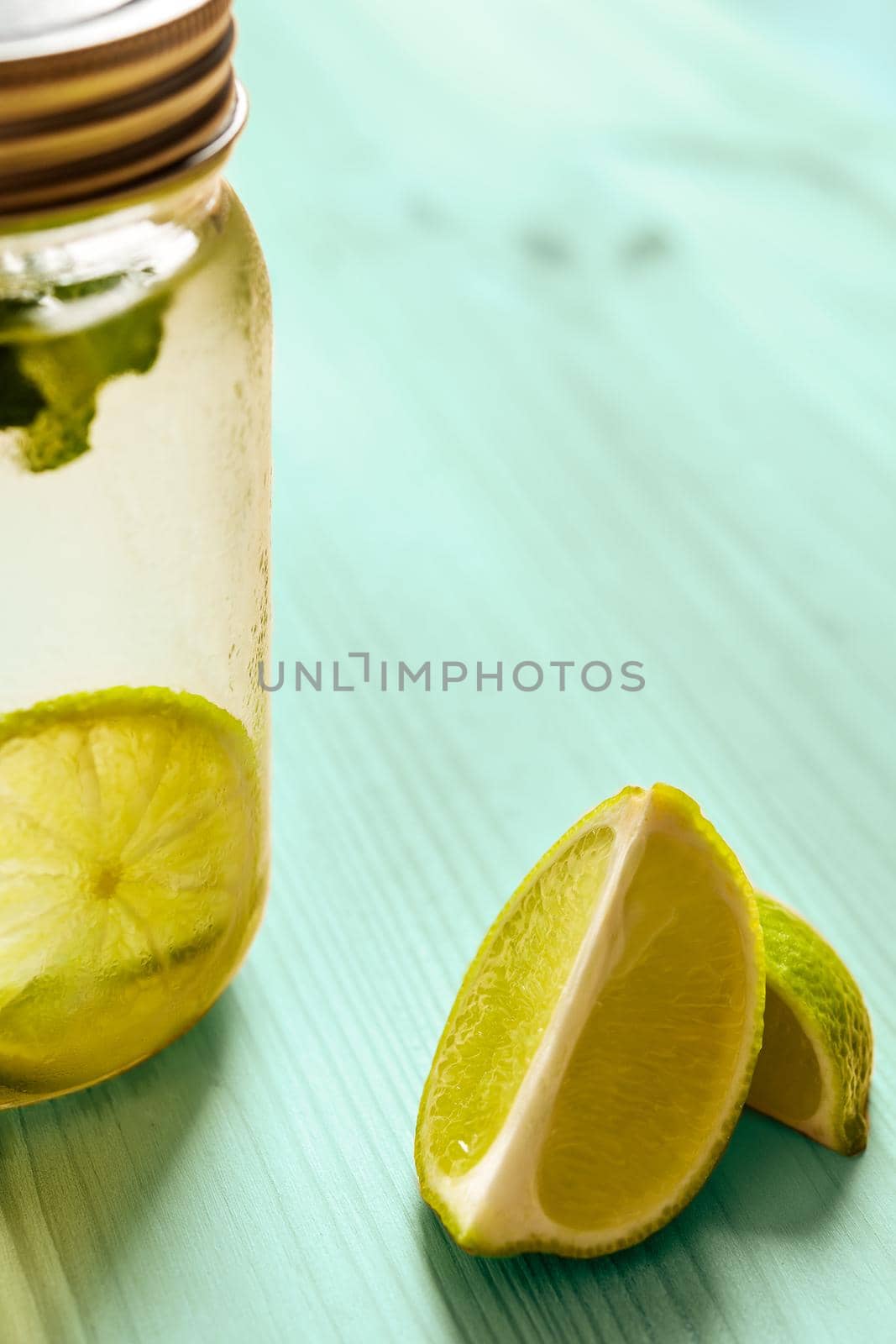 vertical photo of a glass jar with a lid full of lemonade next to some slices of lime, the glass is illuminated by sunlight and on a turquoise wooden table