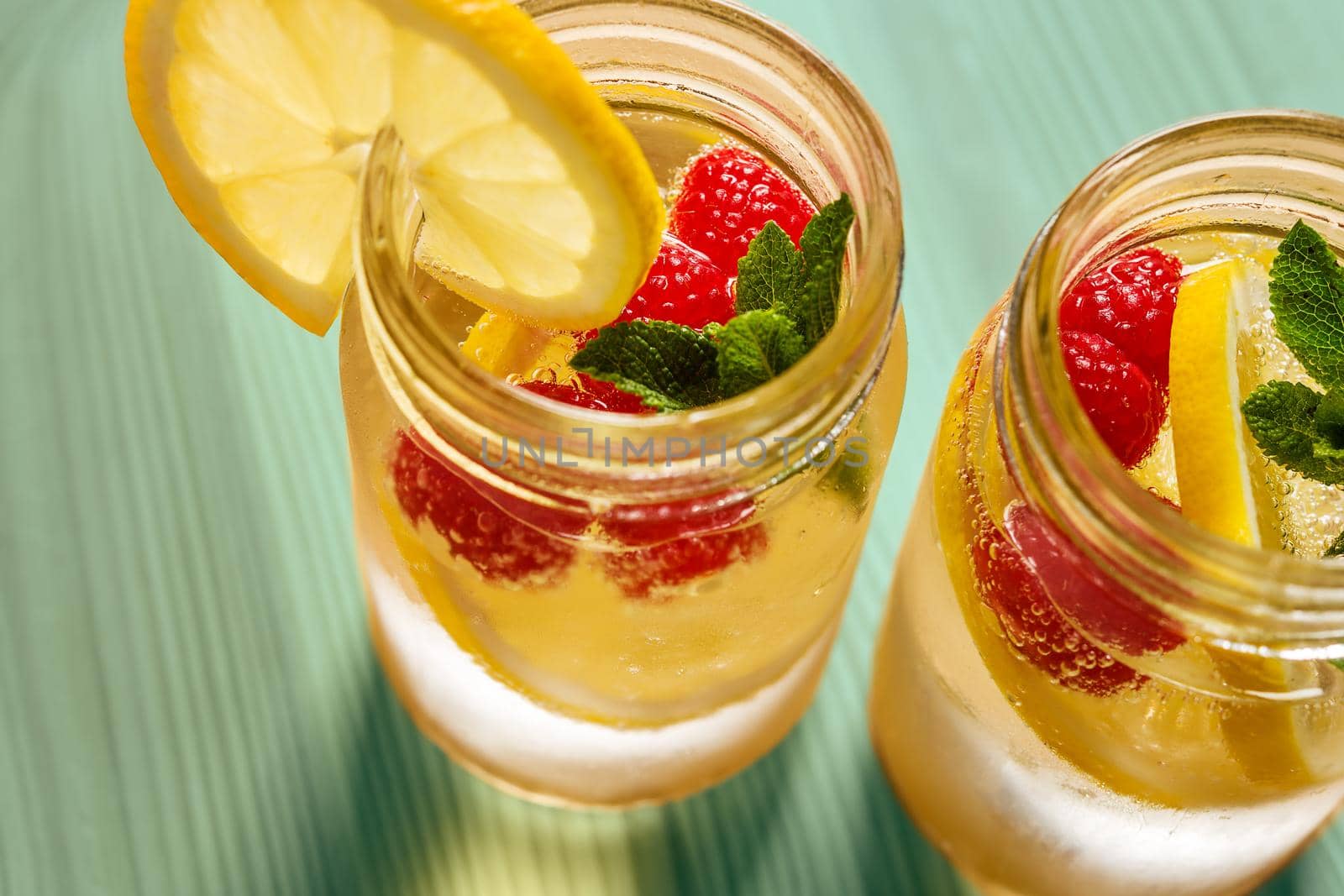 two glass jars with cold water, lemon slices, mint leaves and red berries seen from above, illuminated by sunlight on a green wooden table, citrus summer refreshments background, copy space