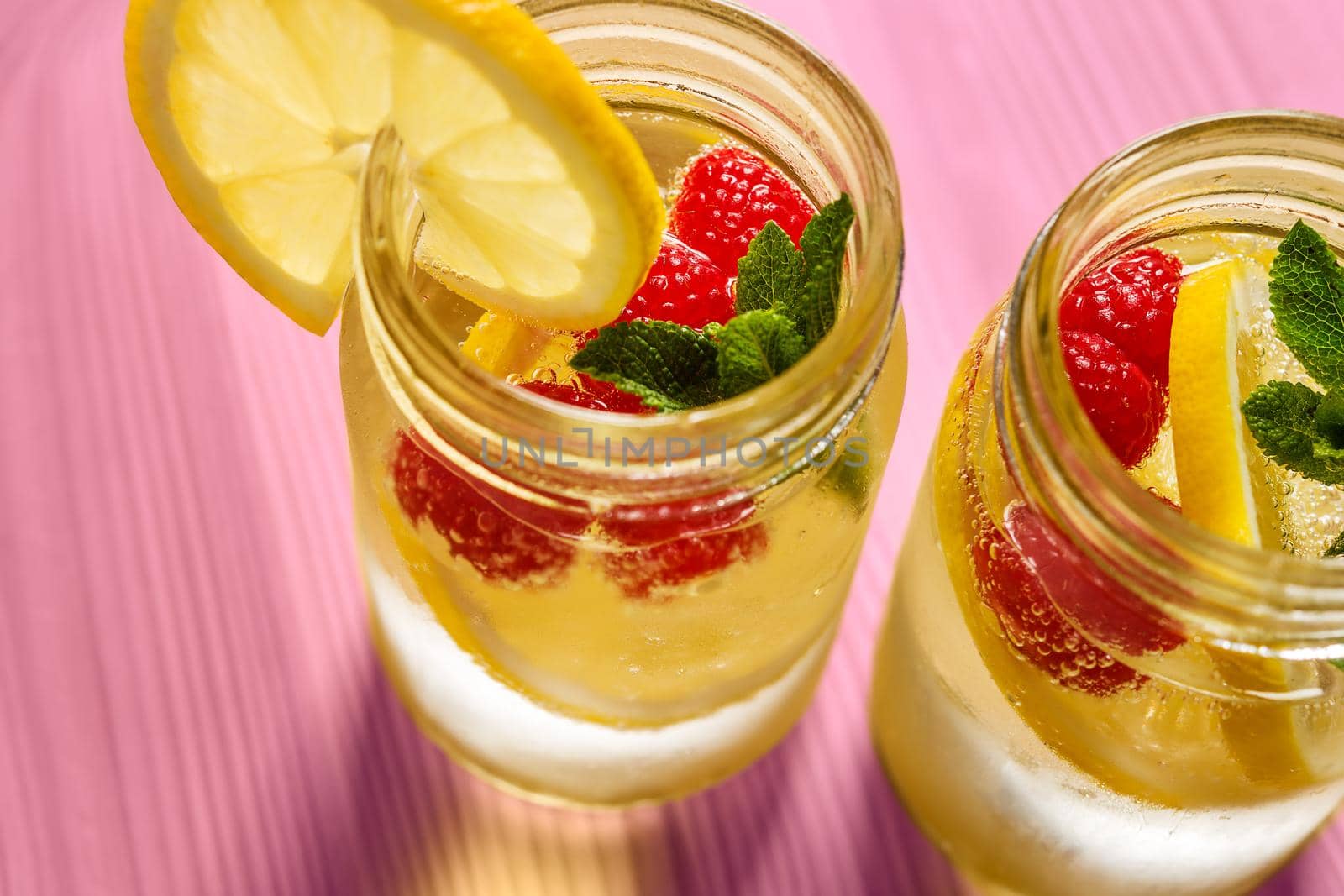 two glass jars with cold water, lemon slices, mint leaves and red berries seen from above, illuminated by sunlight on a pink wooden table, citrus summer refreshments background, copy space