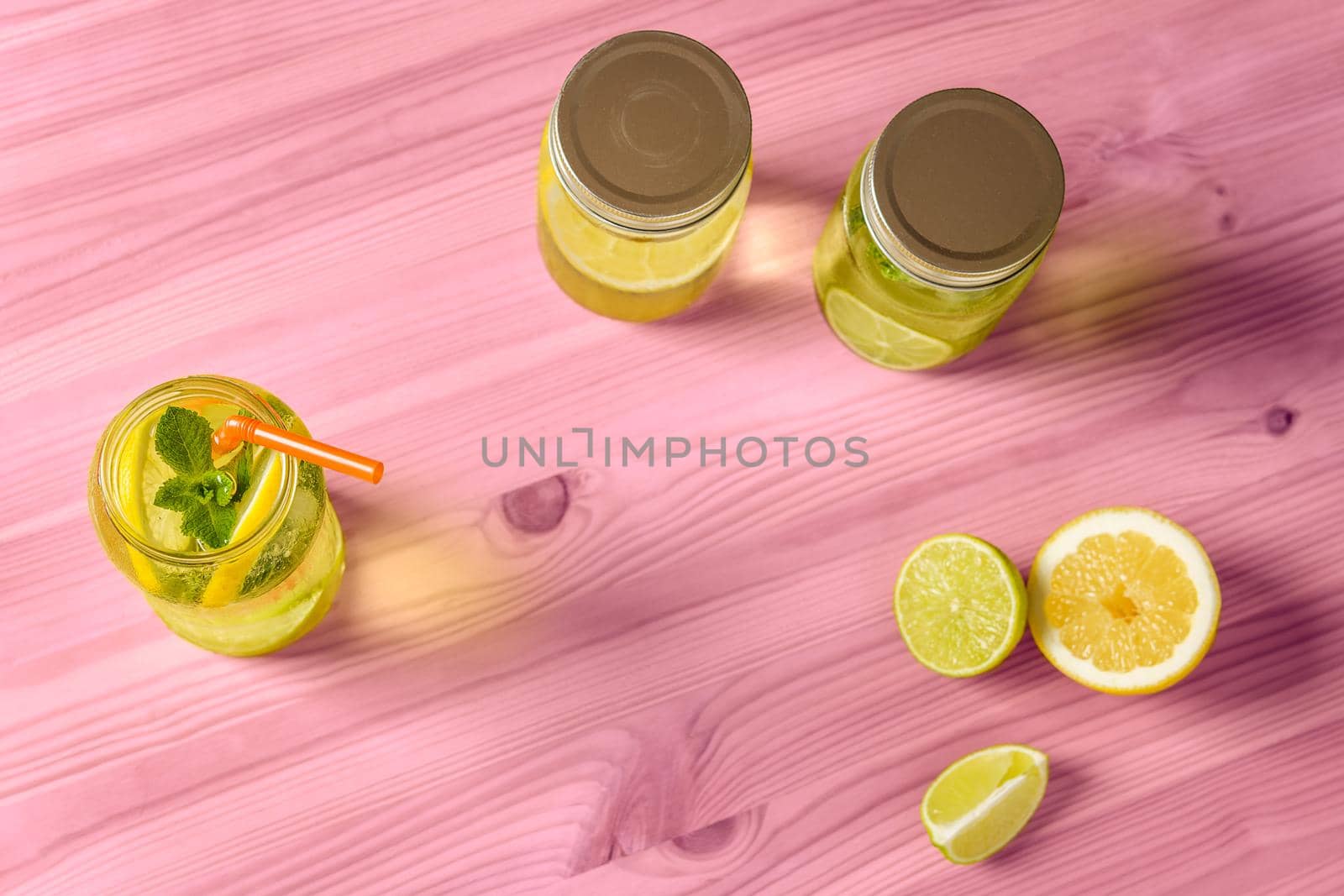 Flat Lay of lemonade with mint and a cane to drink in a glass jar illuminated by sunlight, is on a pink wooden table with two other jars with lid, some pieces of lemon and lime and copy space