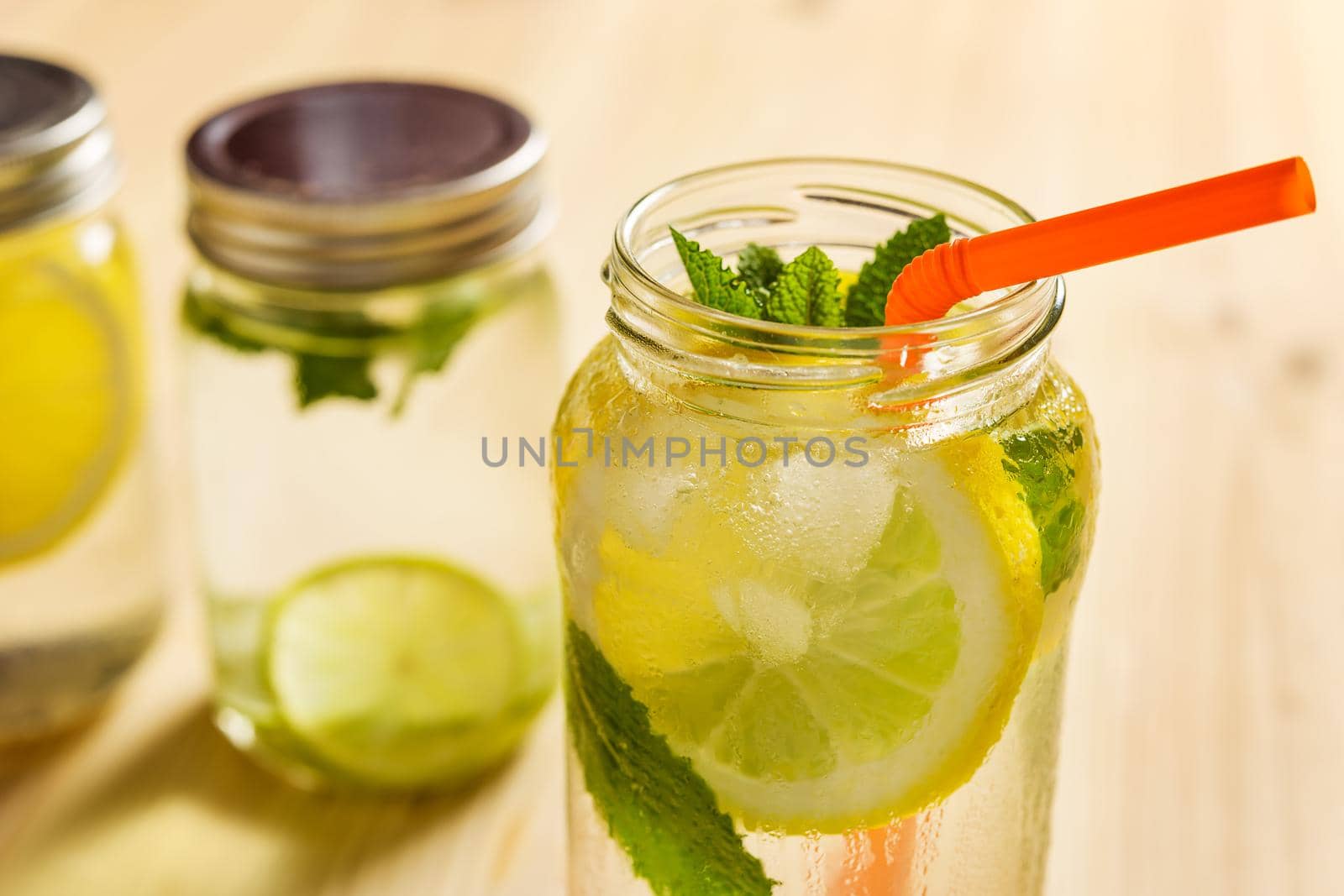 glass jar full of water with ice, slices of lemon and mint leaves, also has a cane to drink and another lemonade unfocused in the background, all is on a wooden table