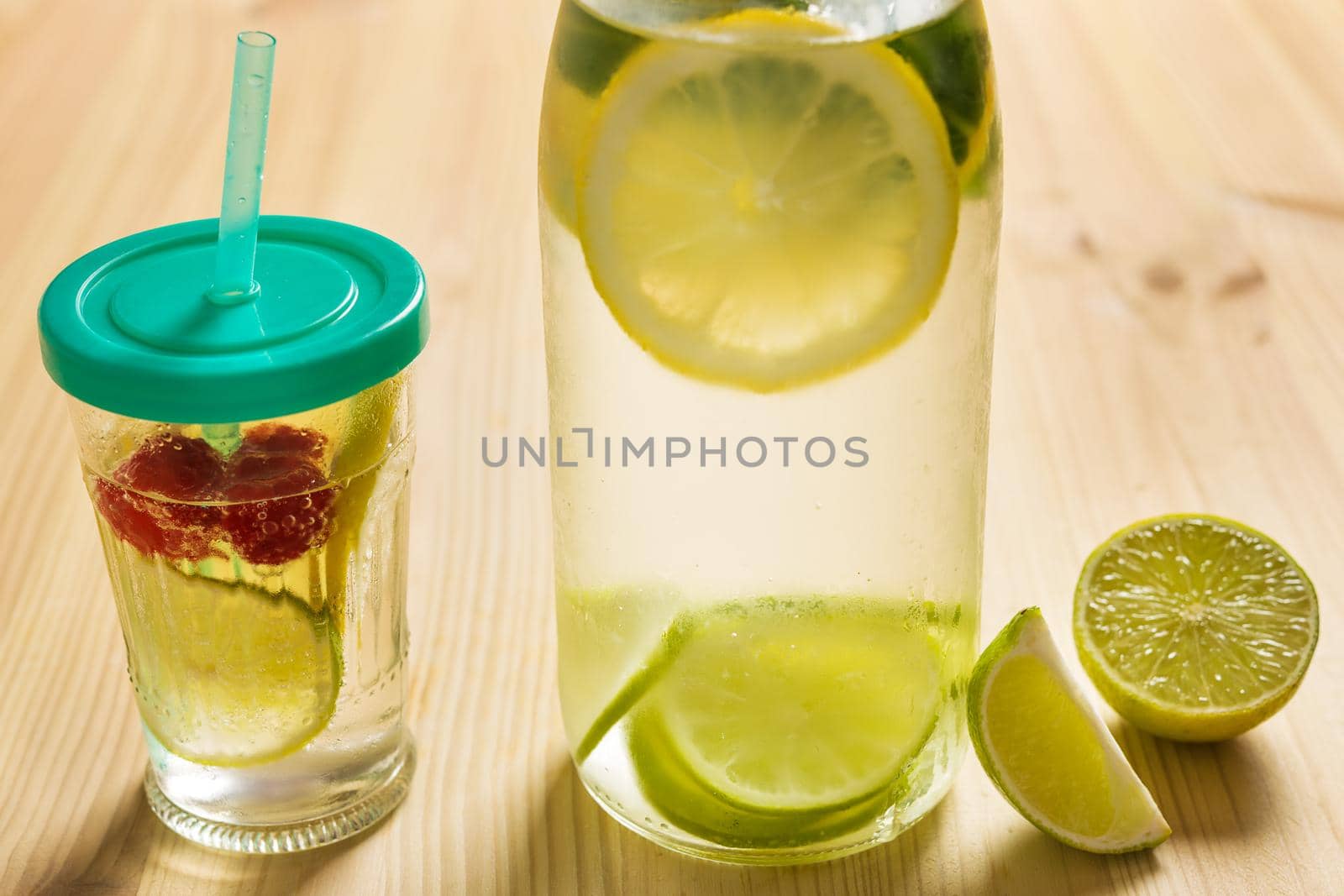 glass bottle and glass with lid and cane, with cold water and slices of lime, lemon, berries and mint, are illuminated by sunlight on a wooden table with some pieces of citrus