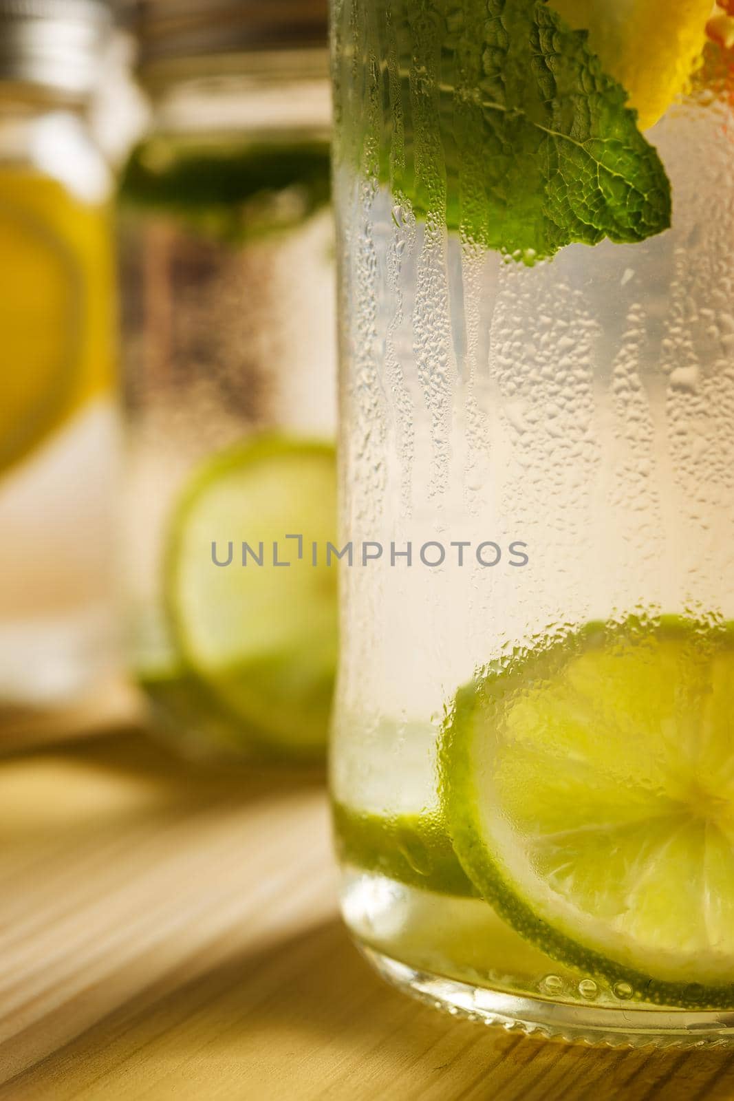 vertical close-up of a glass jar with ice water, lemon, lime and mint leaves on a wooden table and illuminated by sunlight, in the background and out of focus are other lemonades