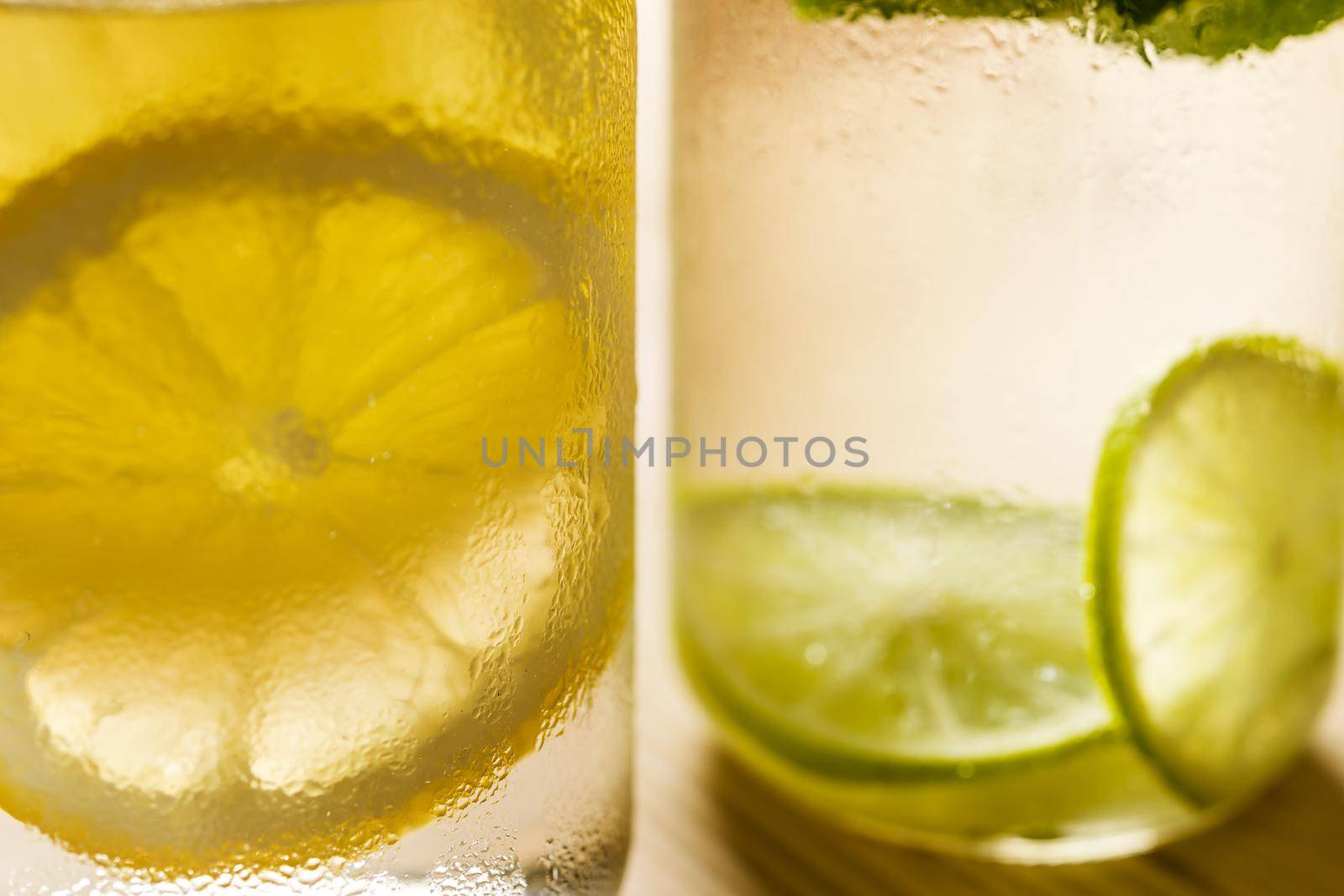 close-up of two glasses with lemonade illuminated by sunlight, one has cold water with slices of lemon and the other with slices of lime and mint leaves