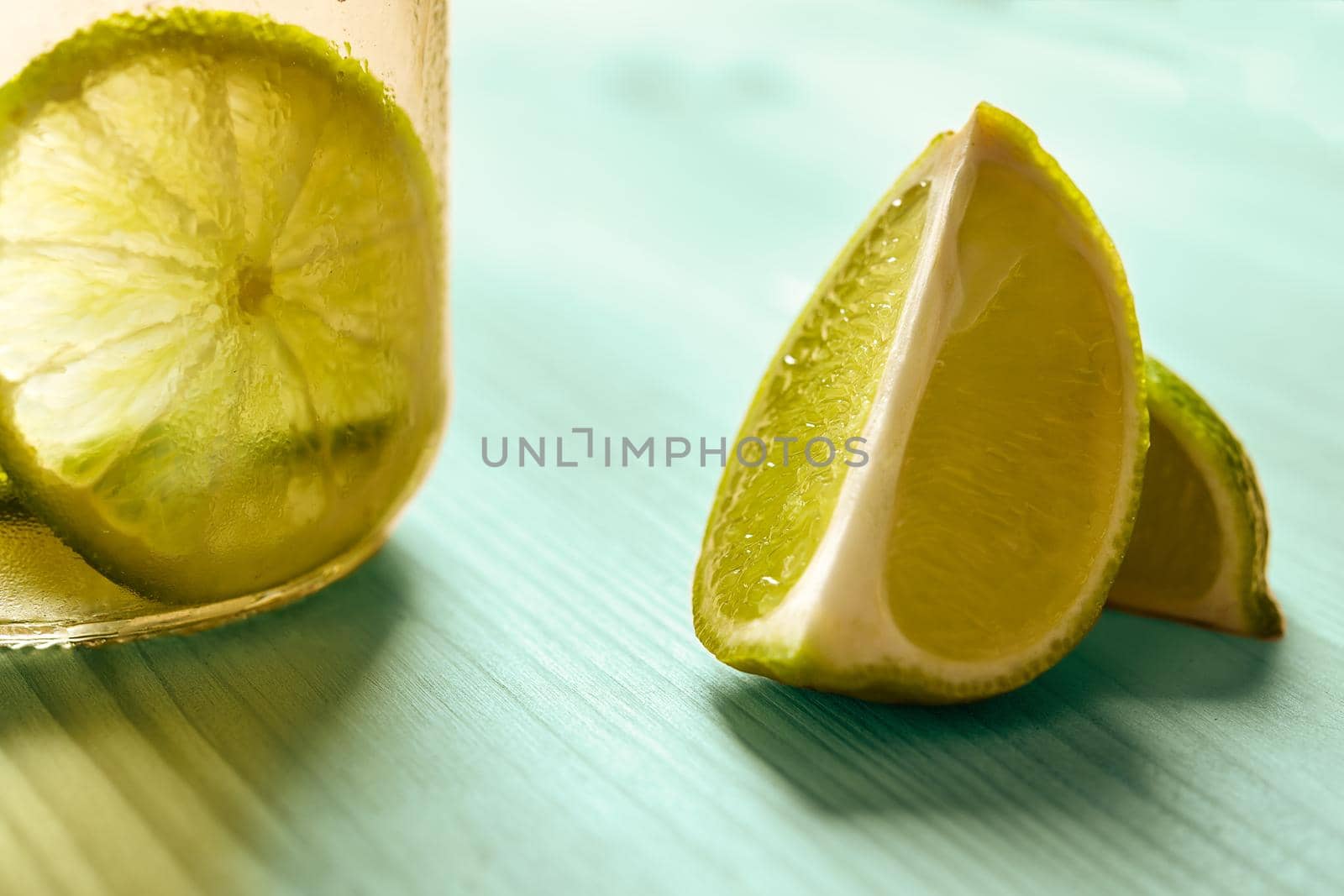 horizontal photo of some slices of lime next to a glass jar with lemonade, this one is illuminated by sunlight and on a turquoise wooden table
