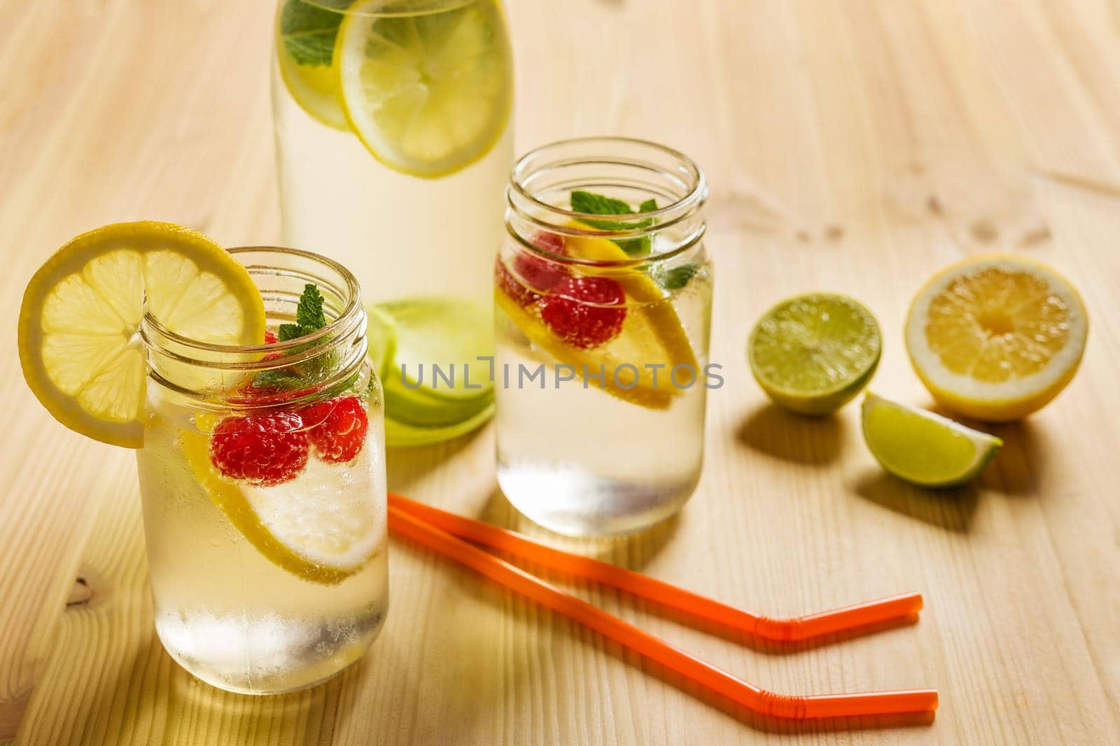 a bottle and two glass jars with cold water, slices of lemon, red berries and mint on a wooden table with canes pieces of citrus lit by sunlight. Summer refreshment background with citrus