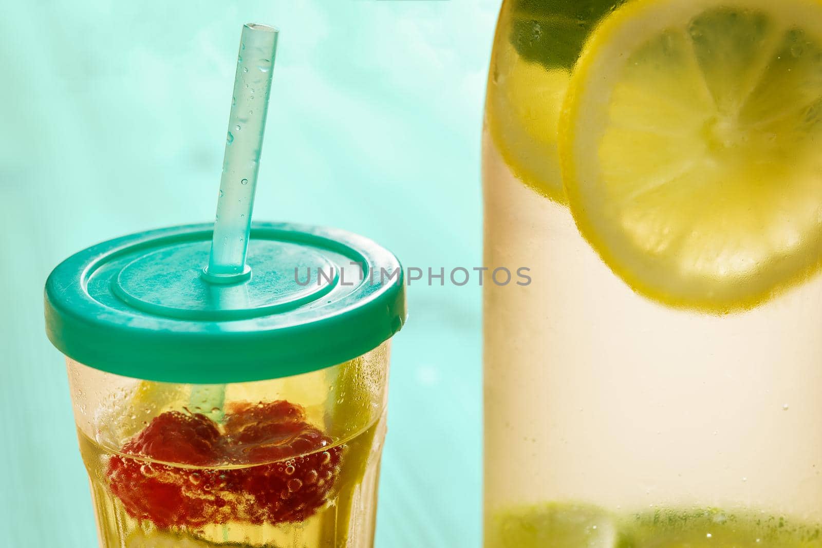 glass with lid and cane and a bottle, with cold water and slices of lemon, lime, berries and mint, are illuminated by sunlight on a wooden green table with some pieces of citrus