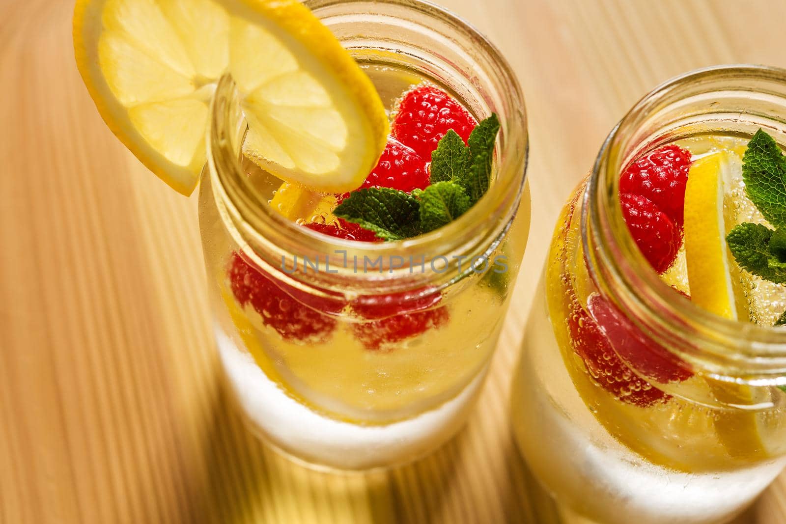 two glass jars with cold water, lemon slices, mint leaves and red berries seen from above, illuminated by sunlight on a wooden table, citrus summer refreshments background, copy space