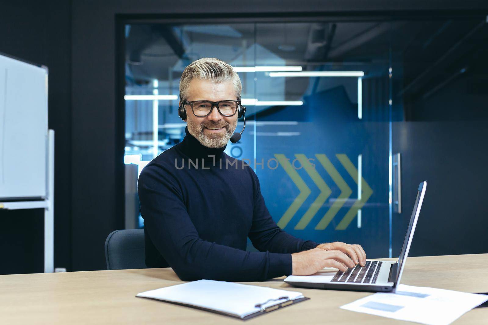 A gray-haired handsome businessman with a headset and a microphone is working on a laptop by voronaman